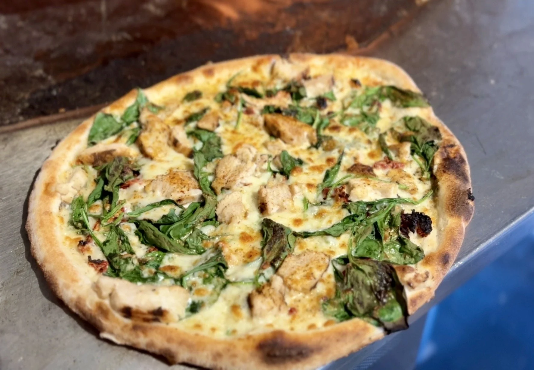 A freshly baked pizza with cheese, grilled chicken pieces, and fresh spinach leaves on a metal tray.