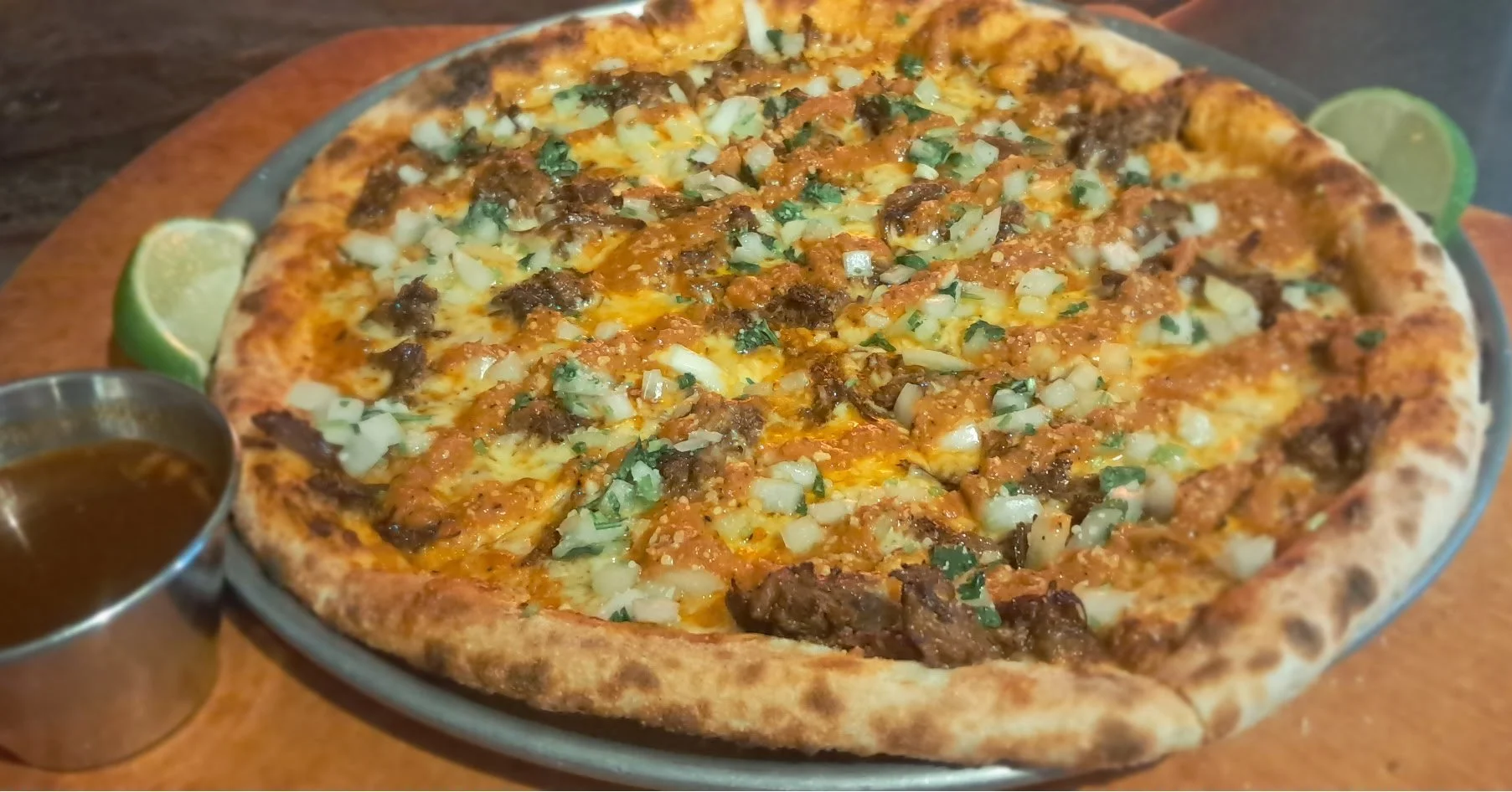 A large, cooked pizza topped with chopped onions, green herbs, beef, and melted cheese on a plate with lime wedges and a small bowl of dipping sauce on the side.