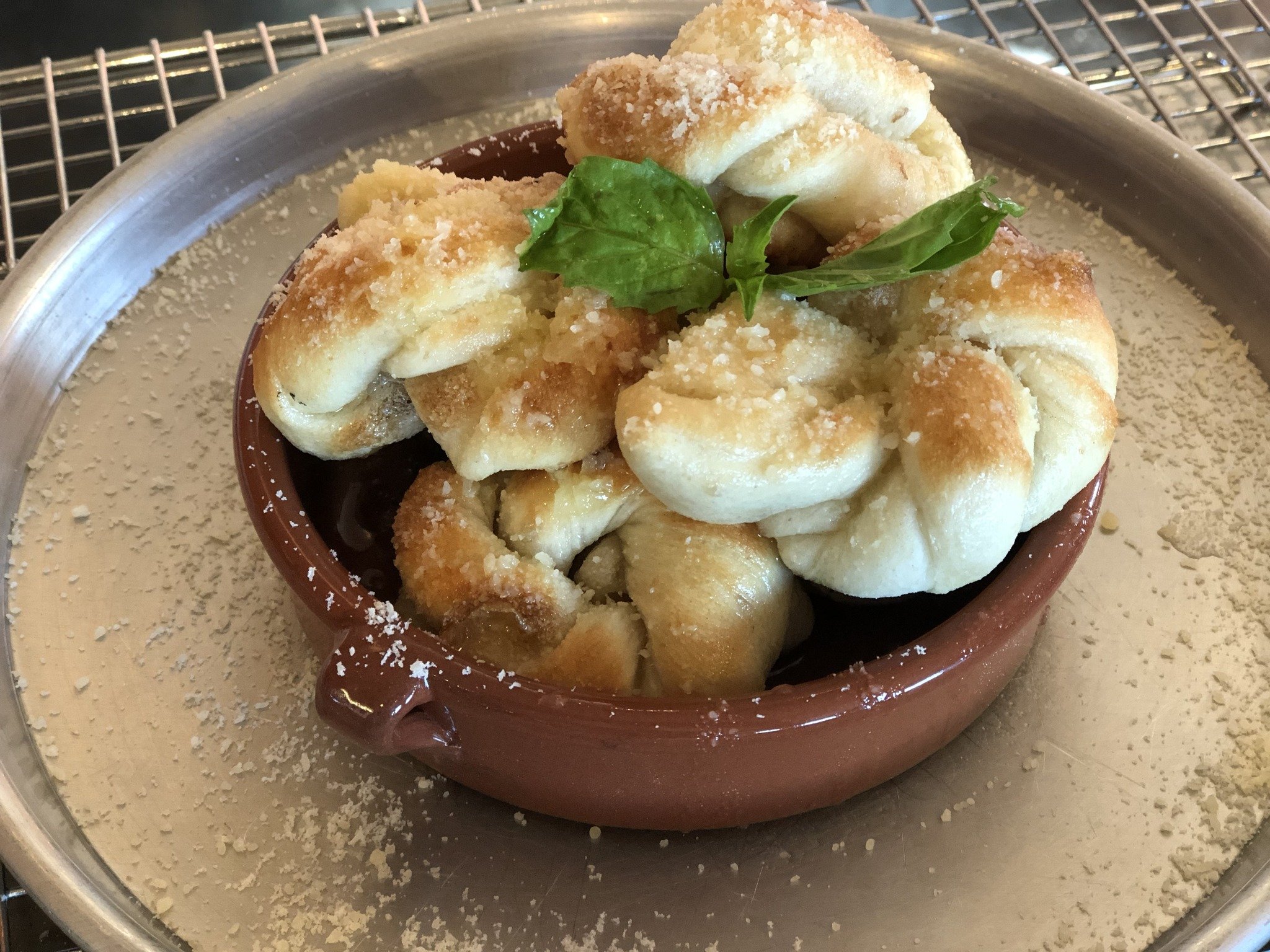 A small brown dish filled with fried dough pieces topped with grated cheese and fresh basil leaves, set on a silver tray with a sprinkle of grated cheese.