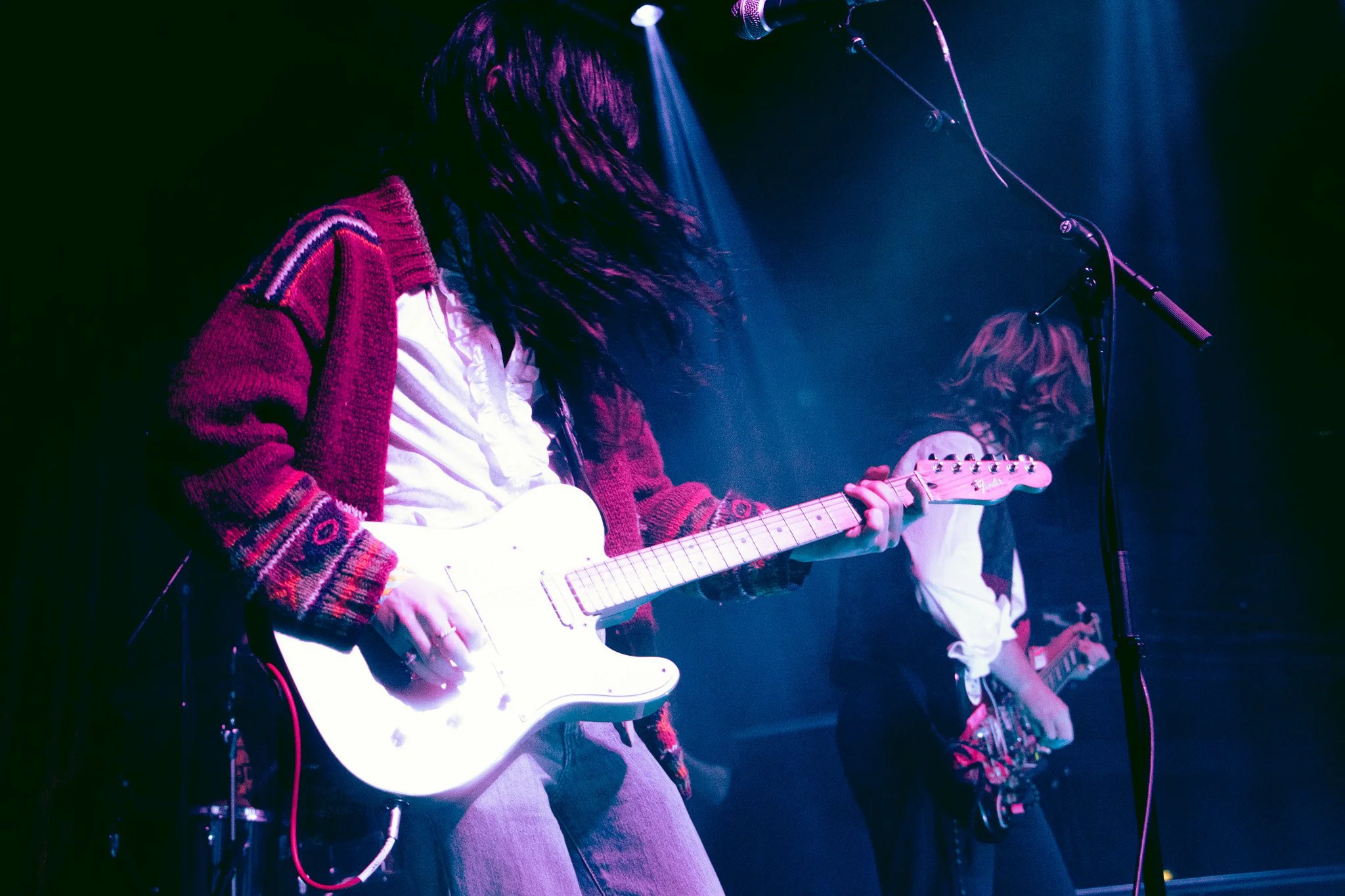 A male musician playing an electric guitar on stage, with long hair covering her face, wearing a colorful cardigan and white shirt, illuminated by stage lights, with another guitarist in the background. The Love Buzz, in The Academy in Ireland. 