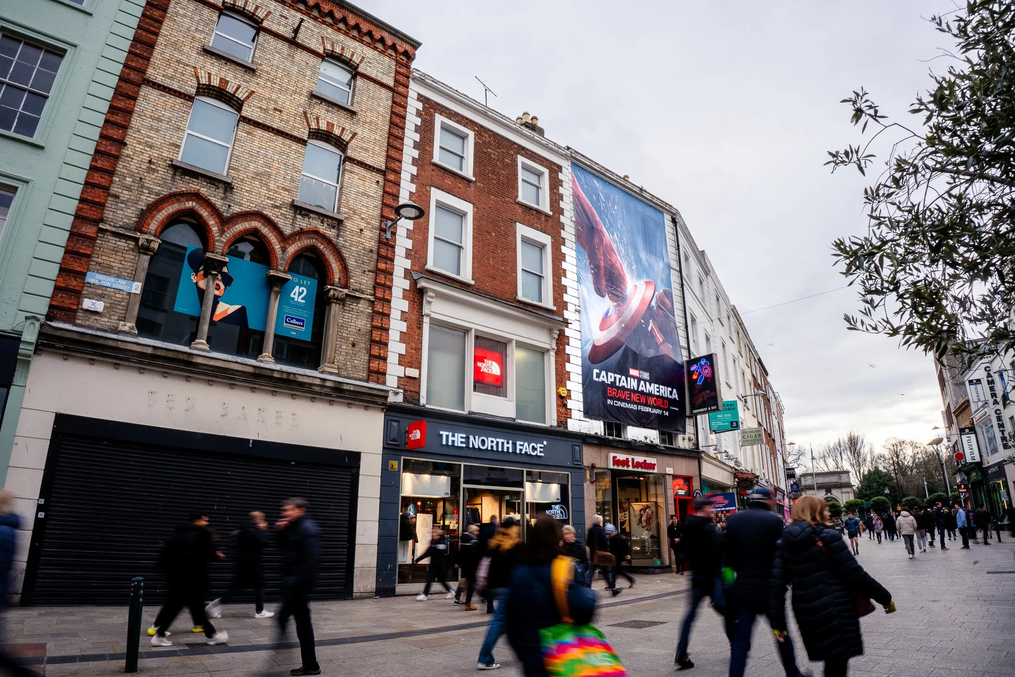 A busy street scene with storefronts including The North Face and Foot Locker, buildings with brick facades, and a large advertisement billboard for the movie Captain America: Brave New World. Pedestrians are walking along the sidewalk under an overcast sky.