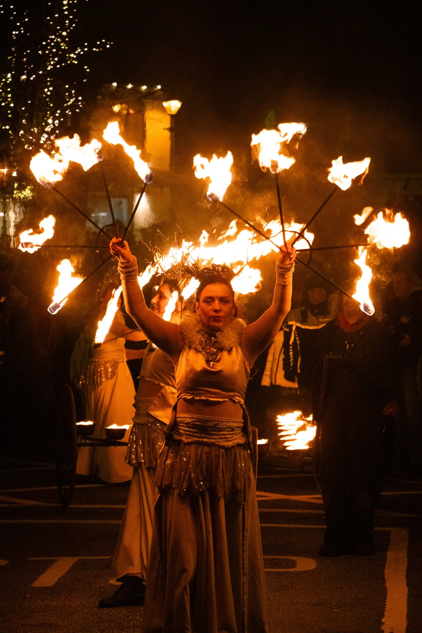 A woman performing a fire juggling act at night during a street event or festival. She is holding multiple flaming torches, with other performers and lights visible in the background. Event Coverage, Shane Lambert, done on St.brigids Day celebrations