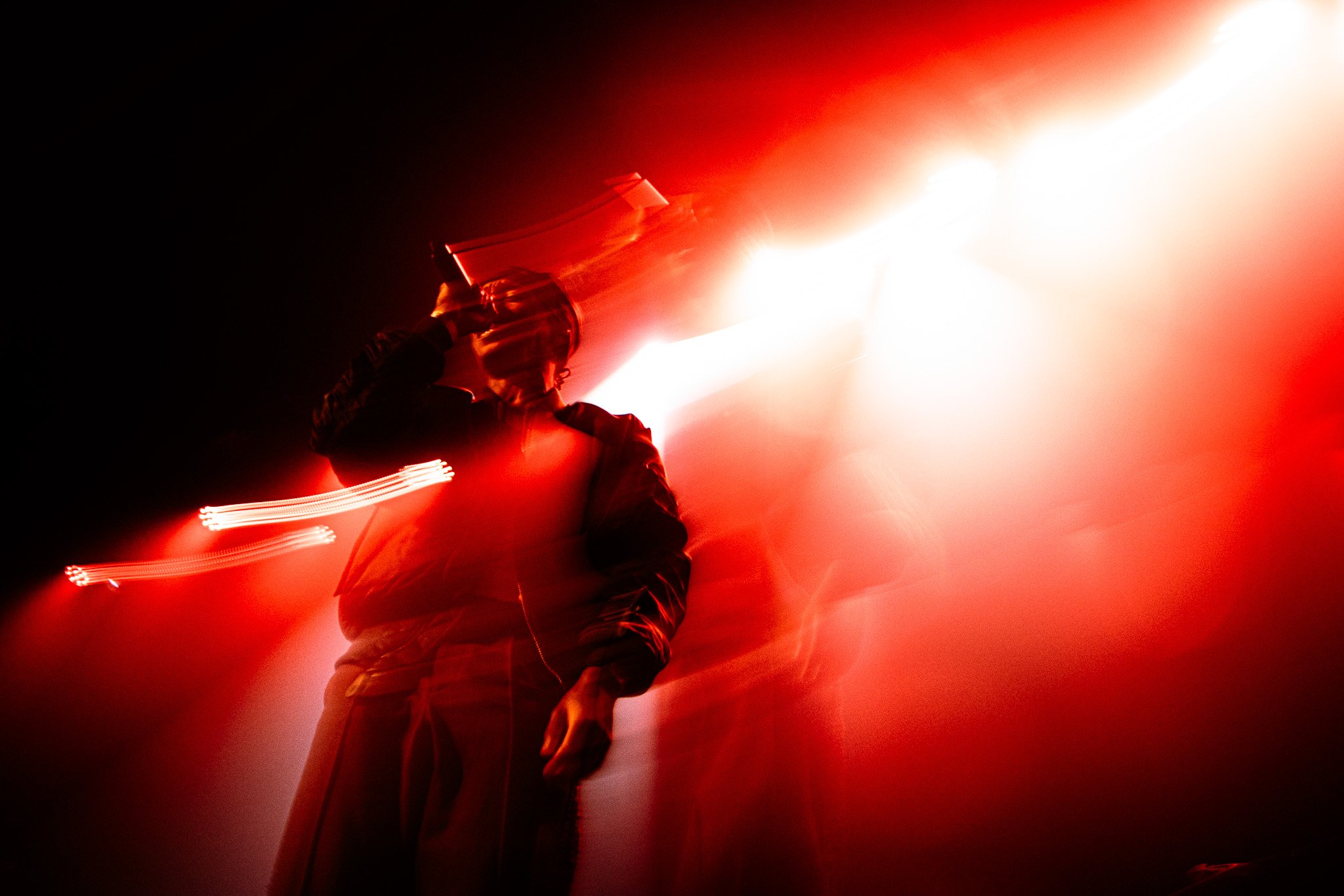 Person holding a bottle to their ear in front of bright light, with red and black tones. Rejjie Snow in the 3Olympia Theatre in 2024. 