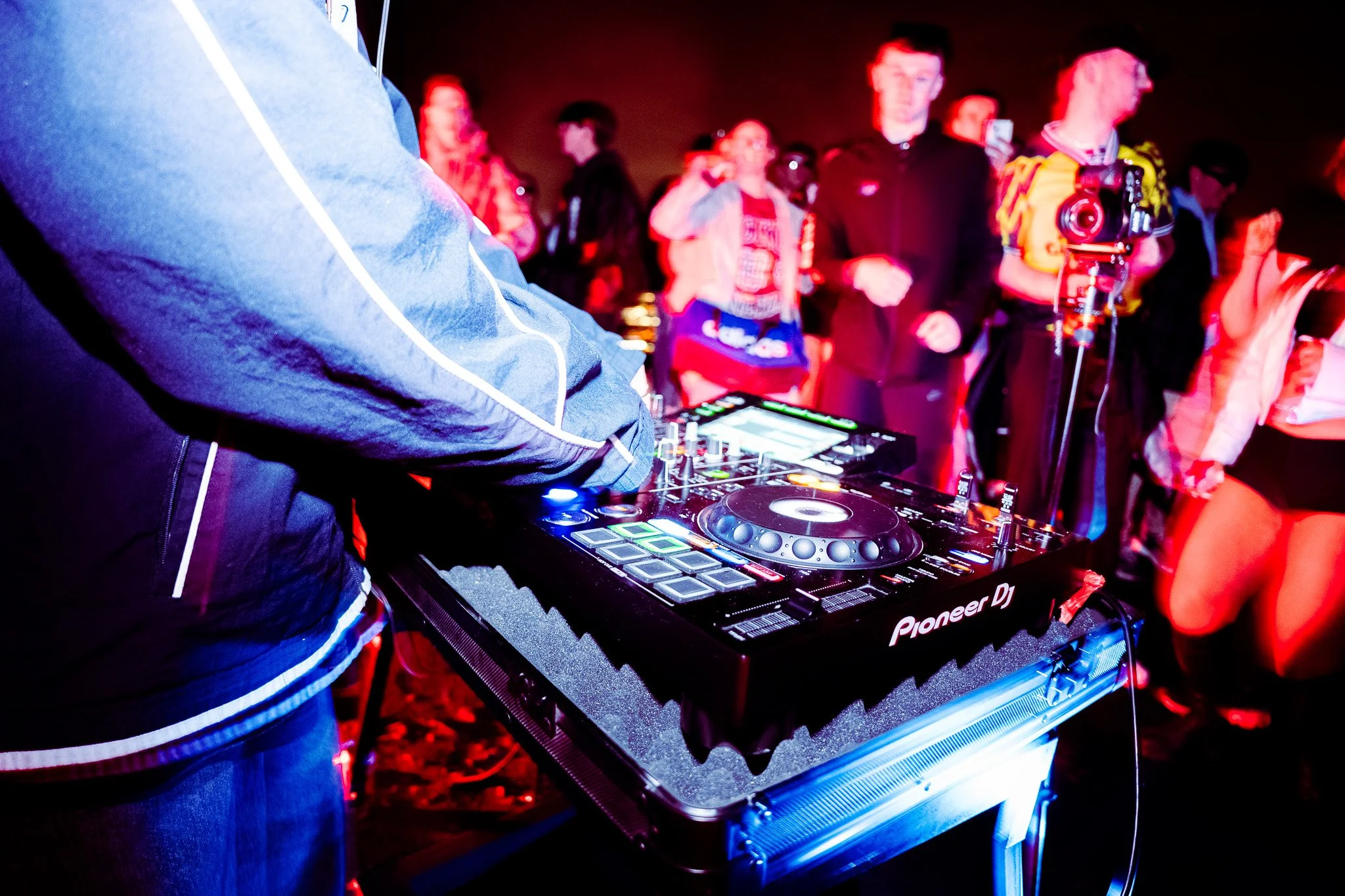 Close-up of a DJ's hand operating a Pioneer DJ controller at a lively party with people dancing and taking photos in the background, illuminated by colorful red and blue lights. Music coverage for irish DJ Childsmind