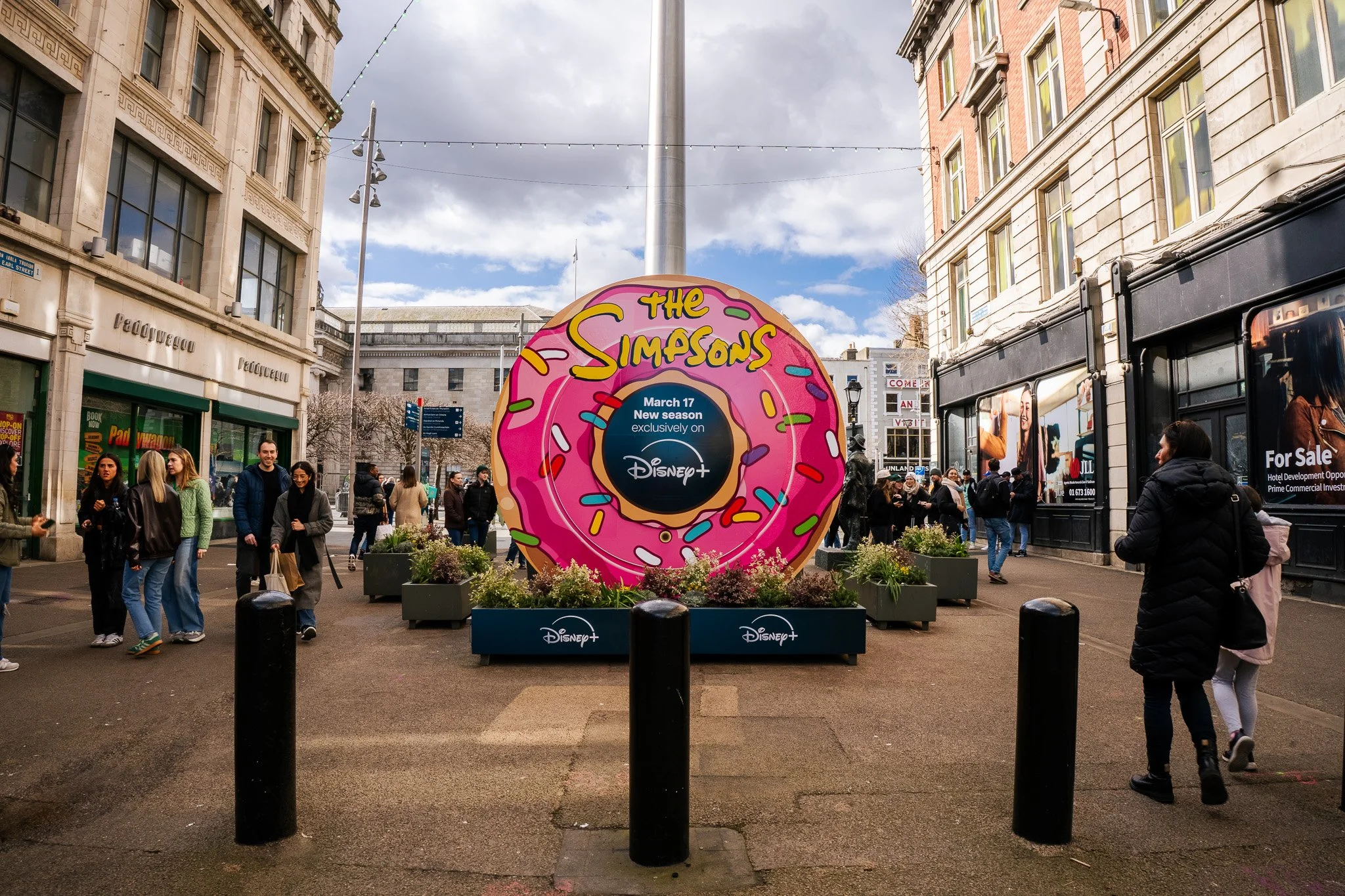 Pedestrian street scene with a large circular billboard featuring a pink donut and decorated with colorful sprinkles, promoting a new season of The Simpsons on Disney+. The street is busy with people walking, shopping, and socializing, surrounded by historic buildings, shops, and billboards.
