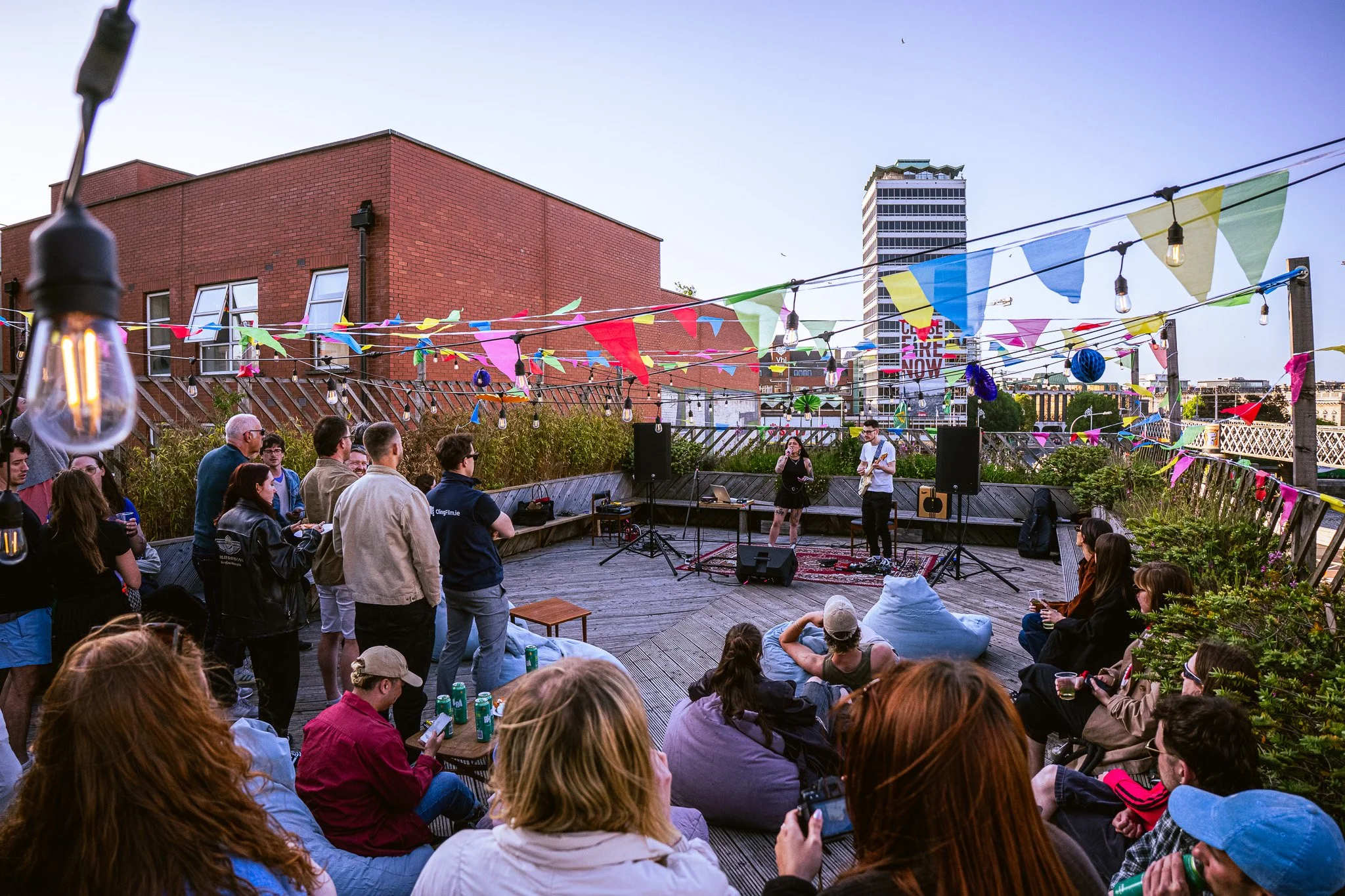 People gathered outdoors on a rooftop deck at sunset, attending a live music event with colorful bunting, hanging lights, and a small stage with musicians. Event Coverage of a musical event on the roof top of the Tara Building. 