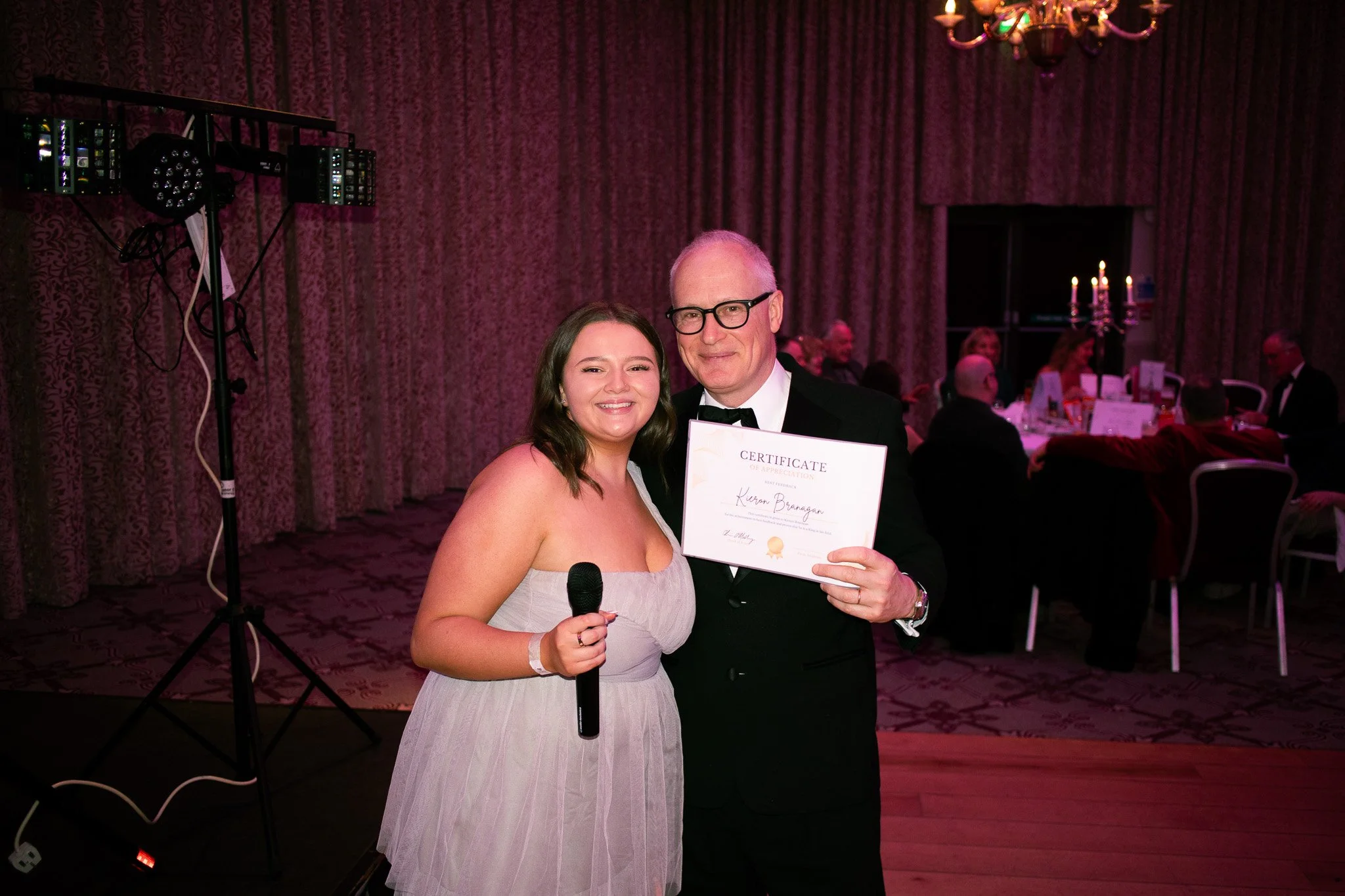 A young woman in a strapless light pink dress holding a microphone smiles next to an older man in a tuxedo holding a certificate. They are at a formal event with a decorated banquet table and people in elegance in the background. Event Photography. 