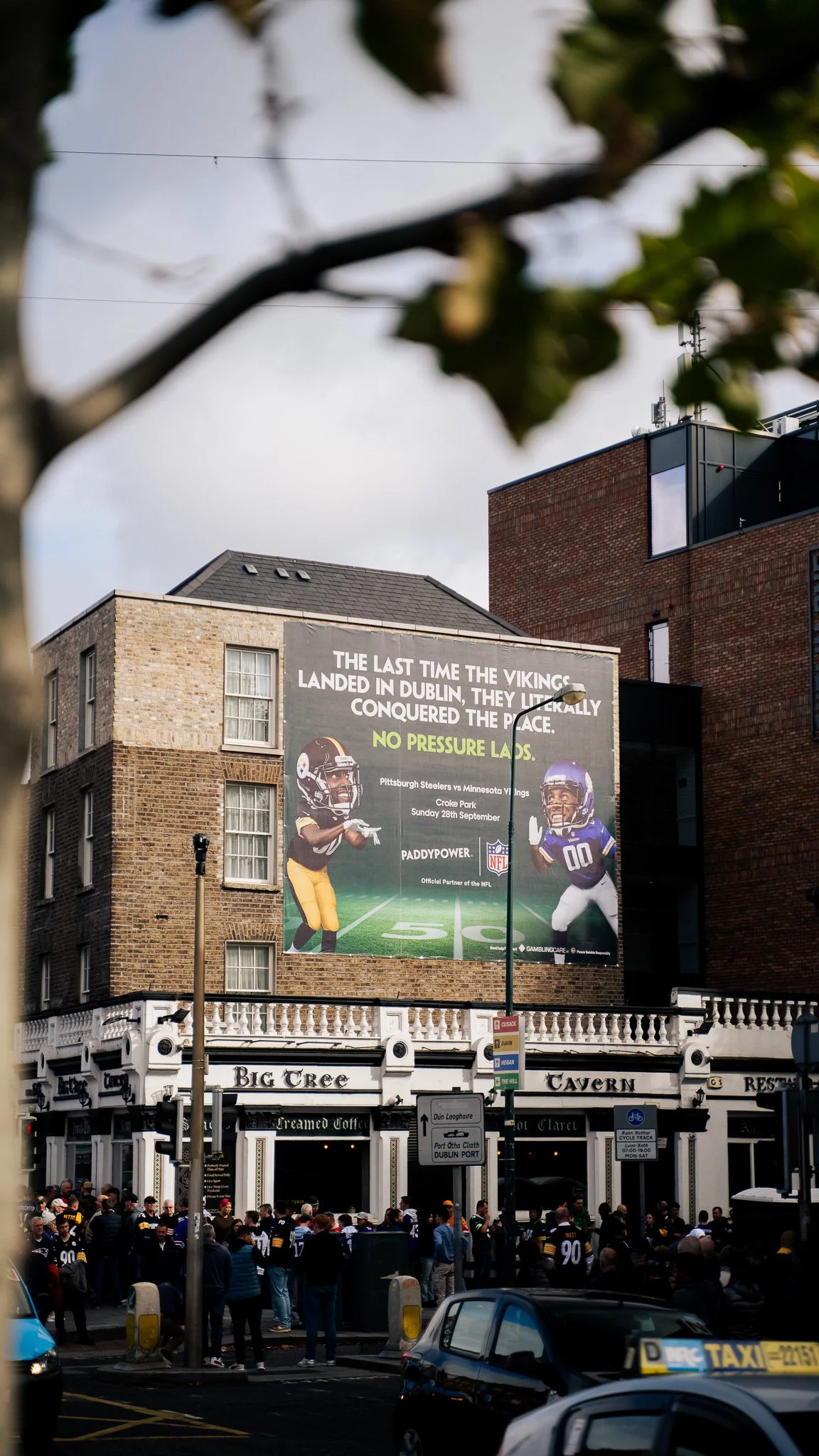 A crowded street scene in Dublin with a large billboard advertising a football game between the Pittsburgh Steelers and Minnesota Vikings, featuring cartoon characters of players, in front of a pub called Big Tree Tavern.