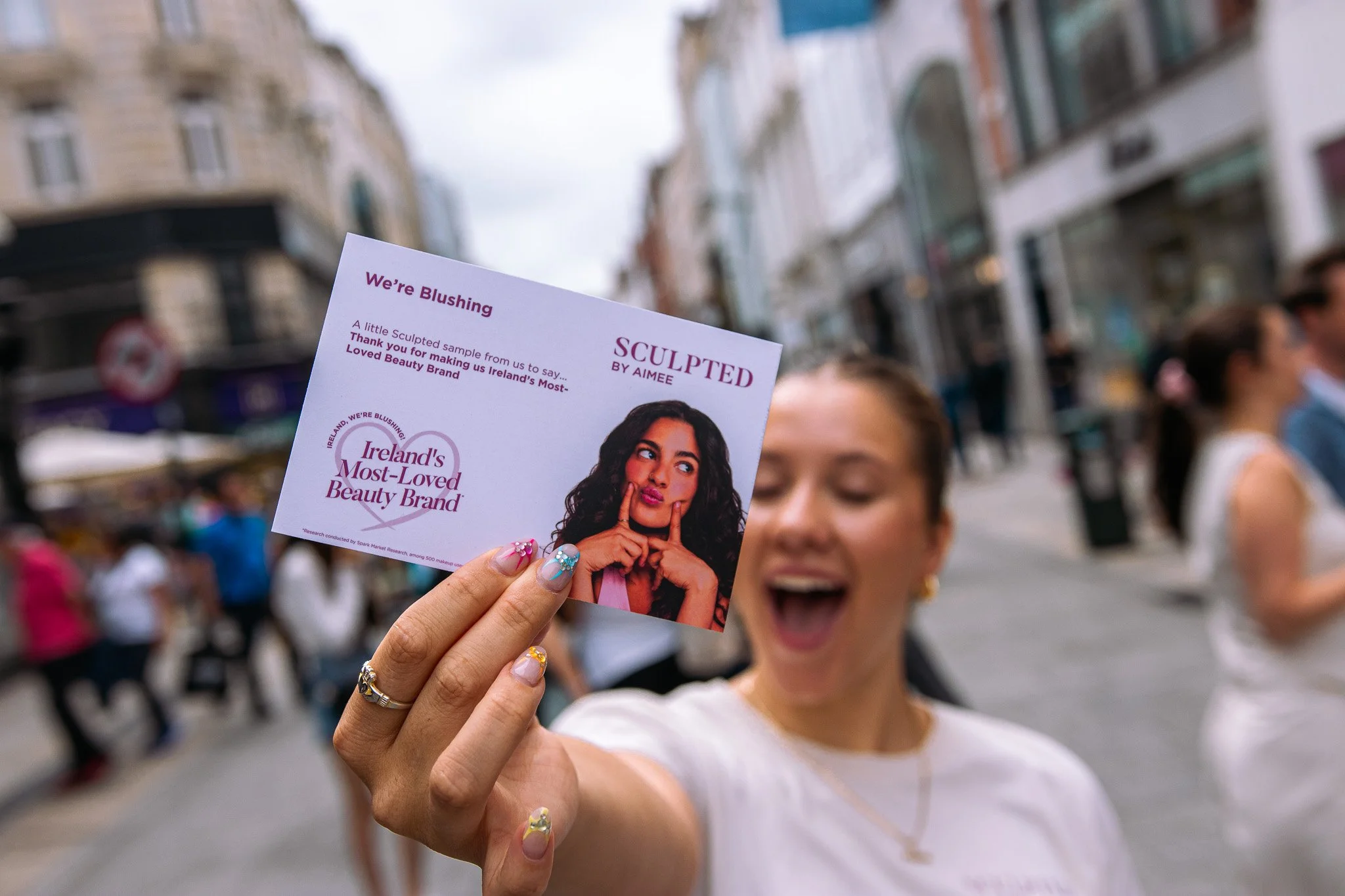 A woman with a big smile holding a promotional card for Ireland's Most-Loved Beauty Brand, with blurred people and city sidewalk in the background.