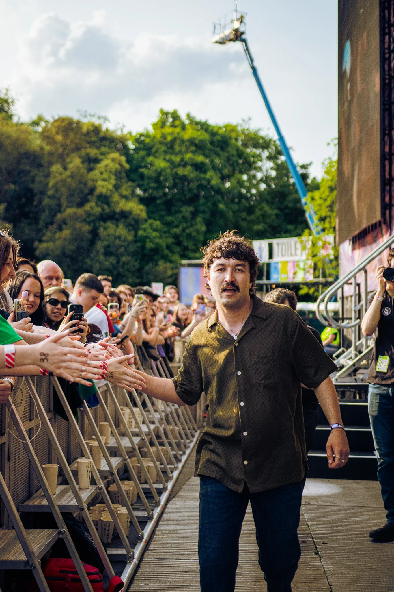 A man with dark curly hair, mustache, and casual attire high-fives fans at an outdoor event with a crowd taking photos and green trees in the background. Gurriers in Dublin, St Annes park, Supporting Irish band Inhaler, 2025. 