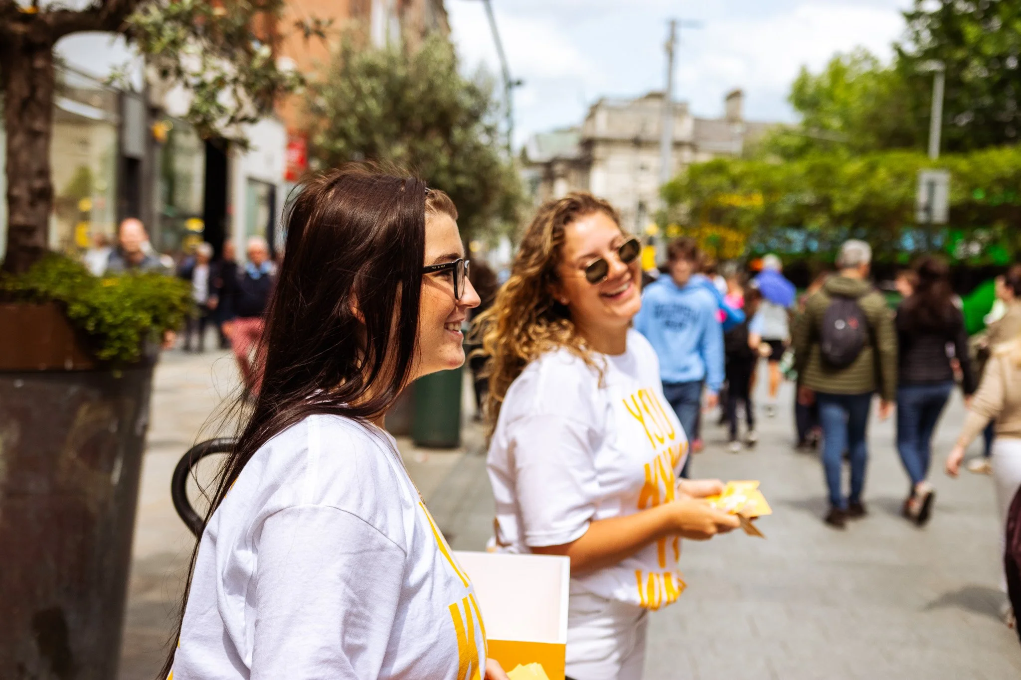 Two young women smiling and talking on a busy city street. One has long dark hair and glasses, the other curly blonde hair and sunglasses. They are wearing matching white T-shirts with yellow text. On Street Brand activation photography coverage. 