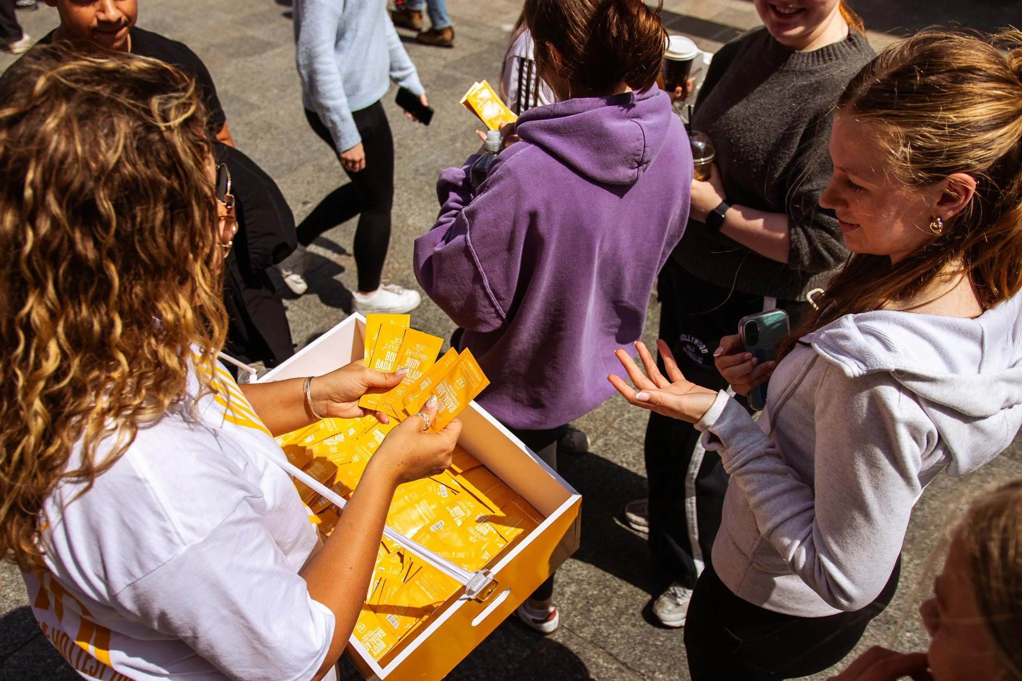 A woman handing out yellow samples to a group of young women outside on a sunny day. On Street Brand activation photography coverage. 