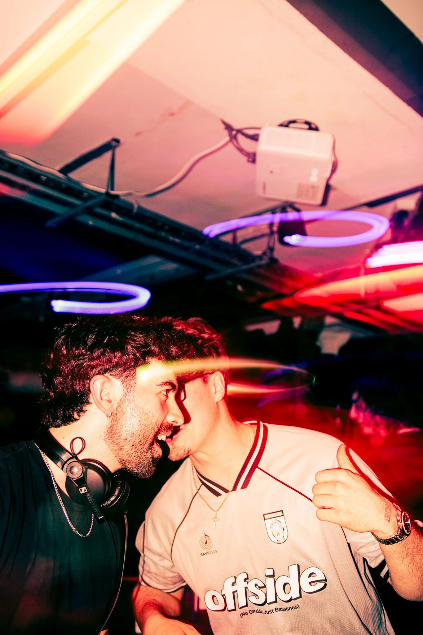 Two men dancing closely in a nightclub with colorful neon lights, one wearing headphones around his neck and the other wearing a white Offside jersey, surrounded by streaks of light. ChildsMind Irish Dj. 