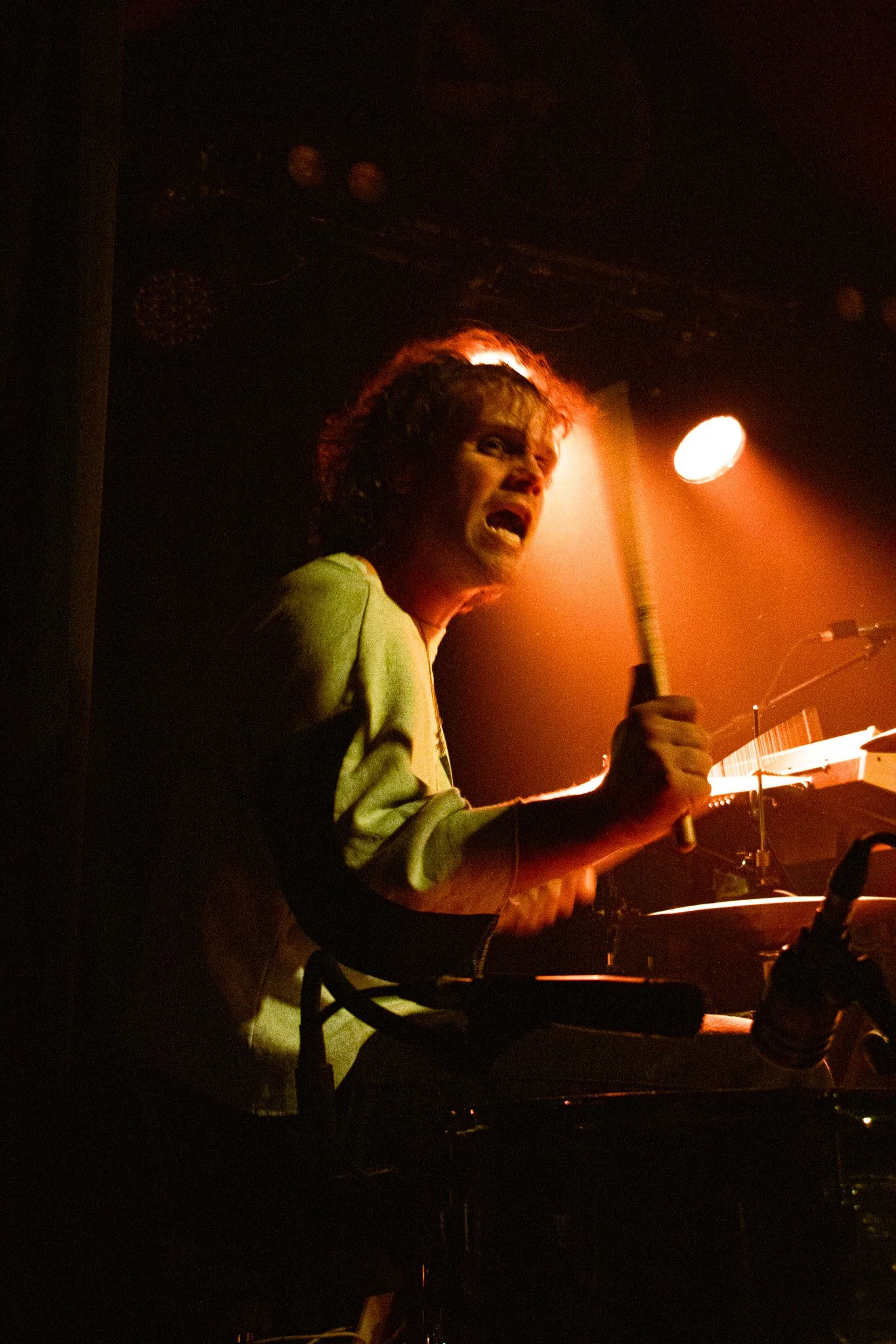 A musician with curly hair and a beard passionately performing on stage, holding a drumstick and playing a keyboard under warm stage lighting. The Grand Social
