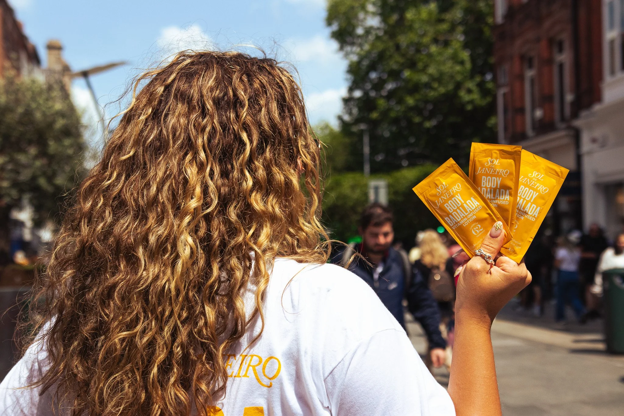 Person with curly blonde hair holding three yellow packets of Sol de Janeiro Body Bad Bolsa in an outdoor urban setting with buildings and other people in the background. On Street brand activation work. 