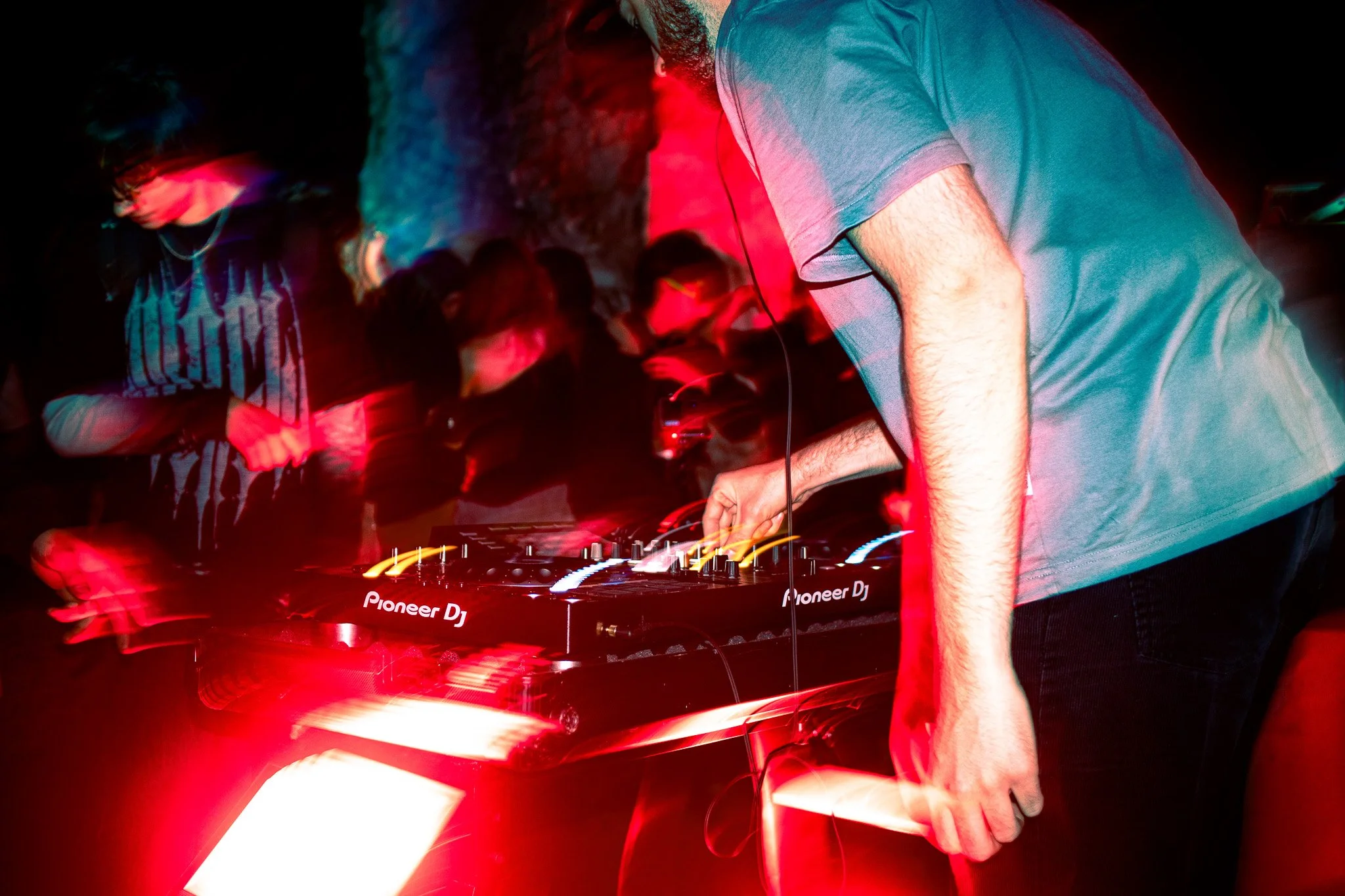 A DJ performs at an outdoor RAVE, surrounded by a crowd dancing, with red and blue lighting creating a dynamic atmosphere.