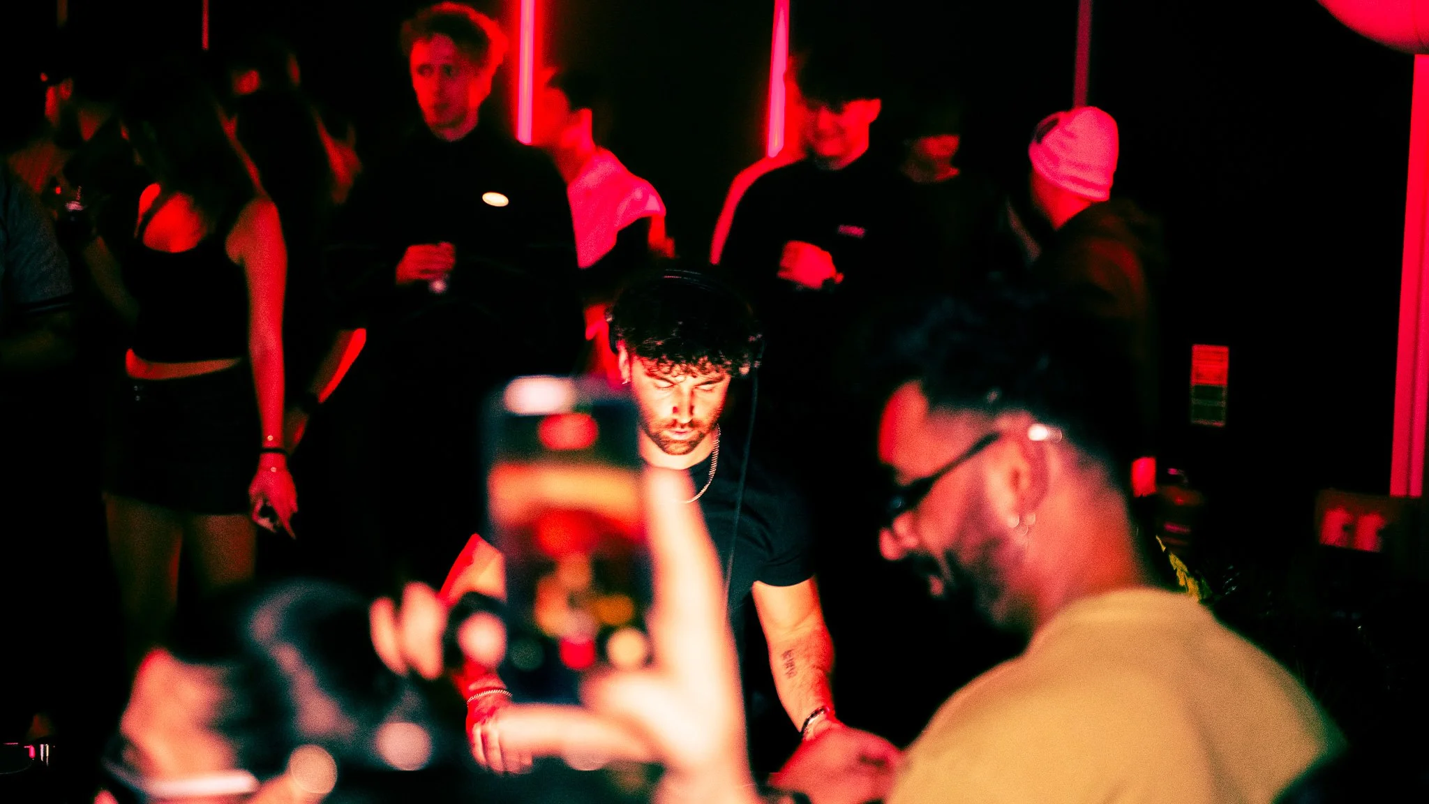 People in a nightclub with red lighting, including a DJ with headphones and glasses, surrounded by others dancing and socializing. Music rave coverage for irish DJ ChildsMind. 
