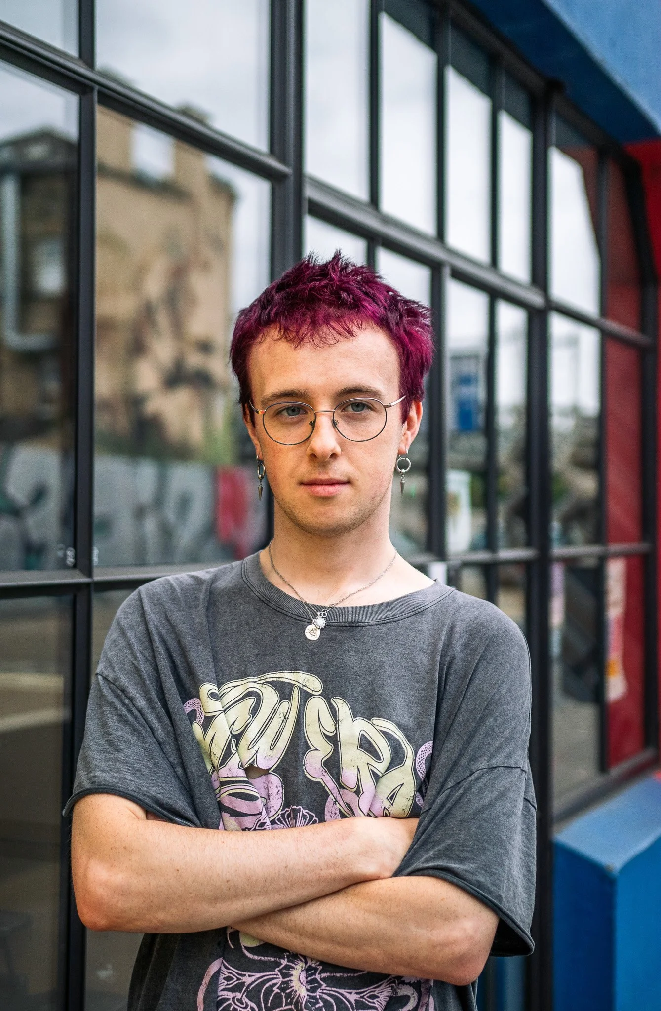 A young person with short purple hair, wearing glasses, earrings, and a graphic t-shirt, standing with arms crossed in front of a windowed building.