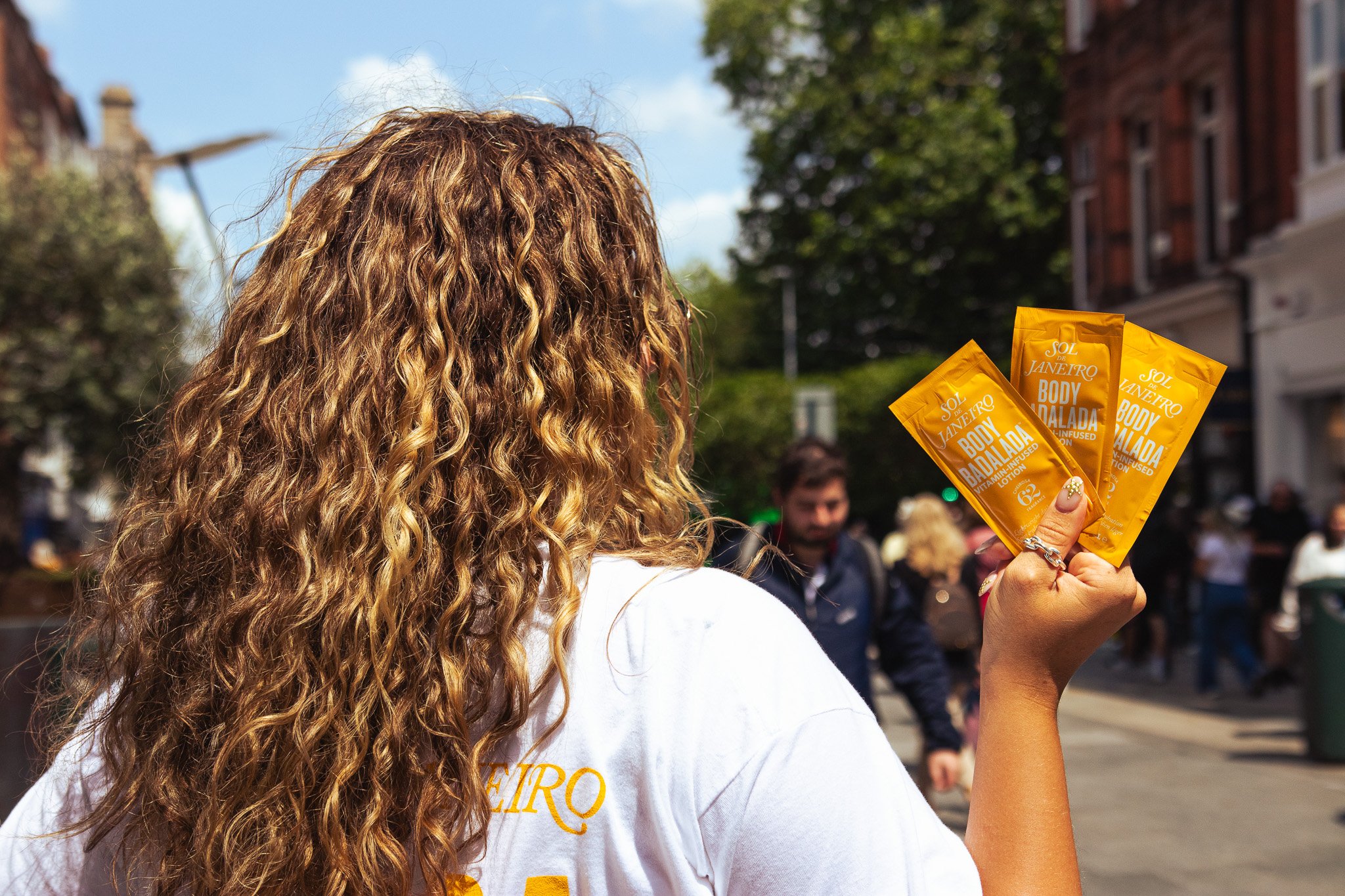 A person with long, curly hair holding three sachets of Sol de Janeiro Body Badalda lotion on a busy city street. On Street Brand activation photography coverage. 