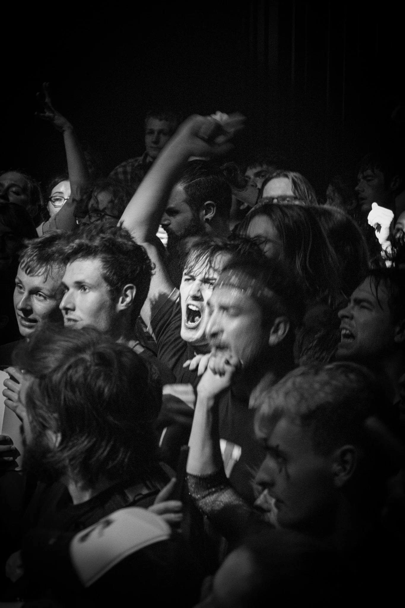 Black and white photo of a crowd at a concert or music event, with some people appear to be singing or shouting, showing intense emotions. Grandson, american musician. 