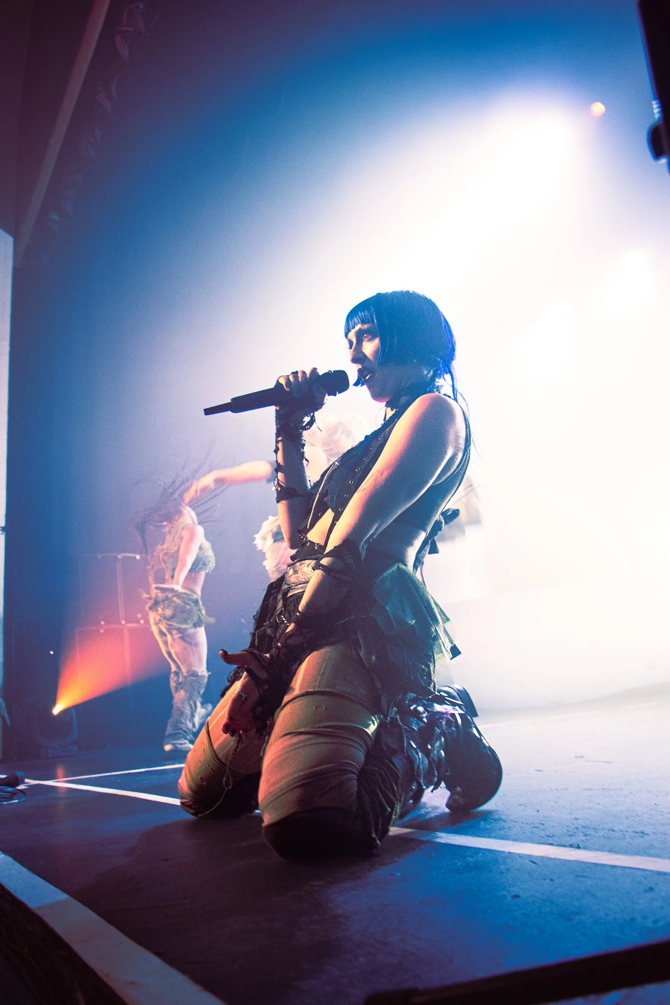 Ashnikko kneeling on stage, holding a microphone, with backup dancers in the background, stage lighting creating a dramatic effect. In Olympia theatre 2023