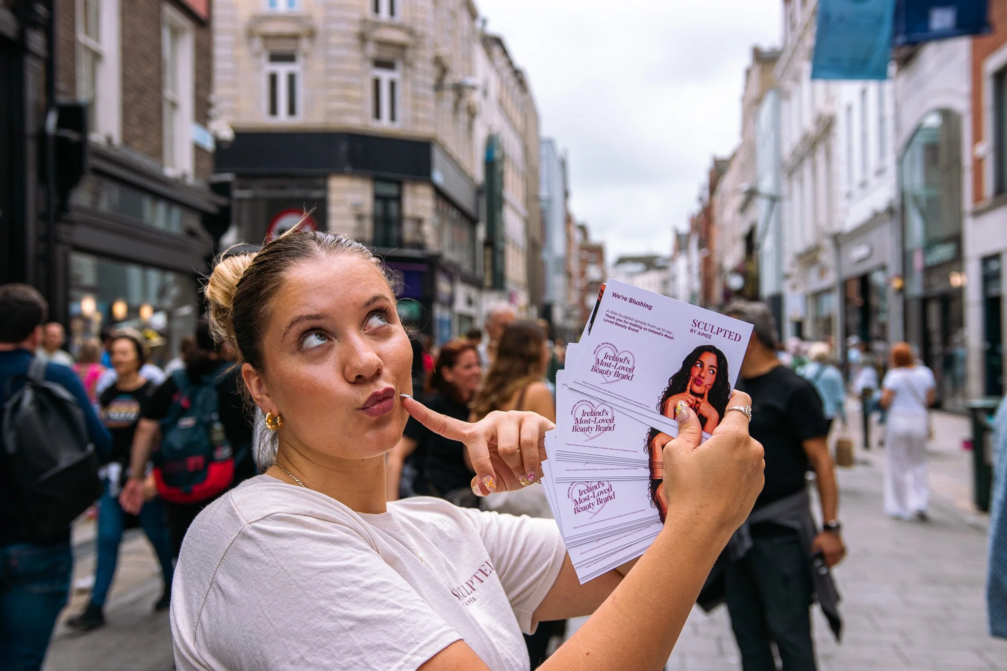 Young woman with blonde hair in a bun holding flyers for Sculpted By Aimee with promotional material in a crowded city street. Client work done, Event Coverage, On Street Activation, Shane Lambert. 