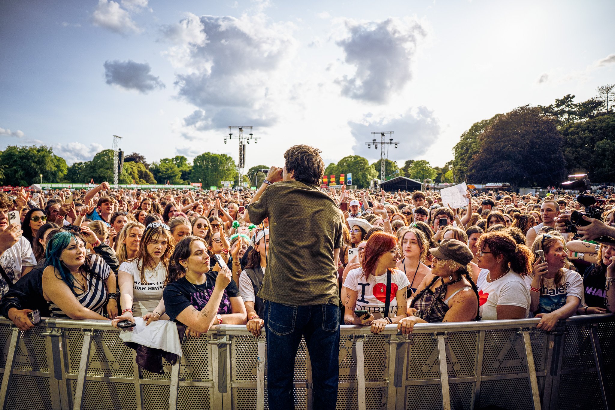 Crowd of concert attendees at an outdoor music festival, with a performer on stage seen from behind, holding a microphone and facing the audience, during daytime with partly cloudy sky. Gurriers playing in St.Annes supporting Inhaler. 