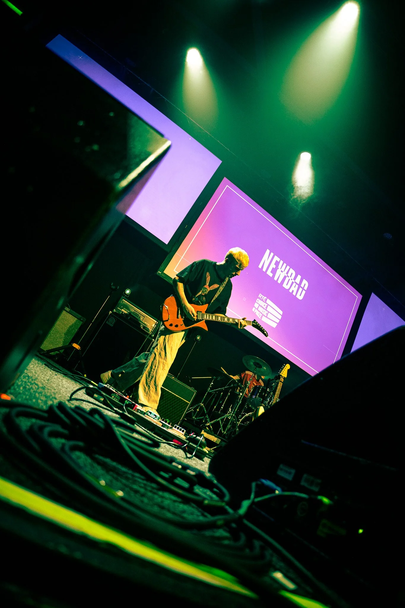 A musician on stage playing an electric guitar during a concert with a large screen behind displaying the word 'NEWDAD' and colorful lighting overhead. Photography coverage at the RTE Music Choice Awards. 