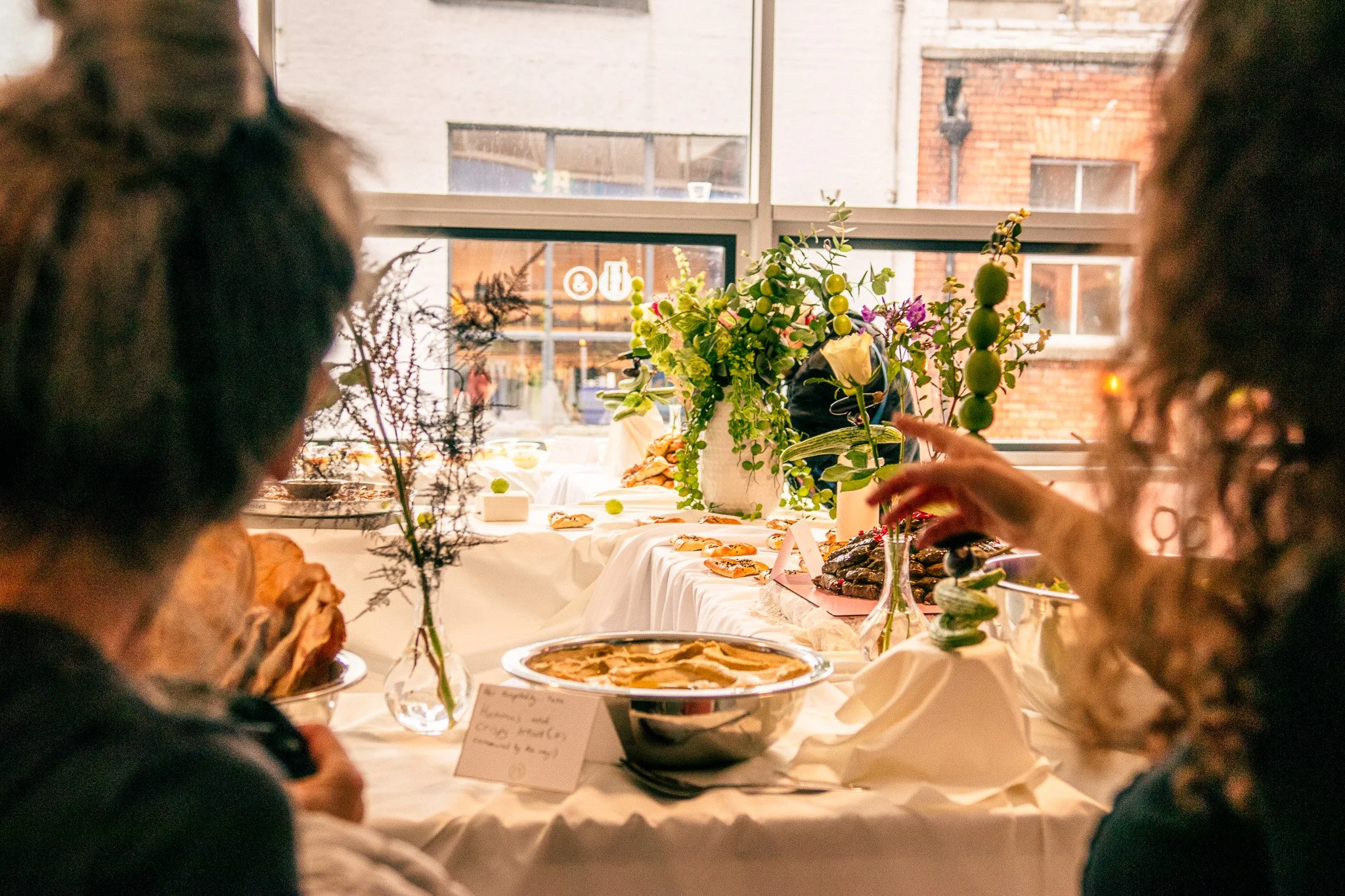 People enjoying a buffet table with a white tablecloth, decorated with flowers and various dishes, in front of a window showing buildings outside. Event Coverage done for Tawla Collective in the Project Arts Centre. 