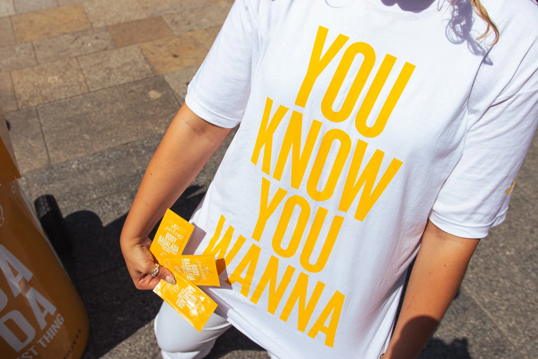 A person wearing a white t-shirt with yellow text that reads 'YOU KNOW YOU WINNA'. They are holding multiple yellow packets in their hand, which appear to be promotional items. The person is standing on a sidewalk with paved stones.