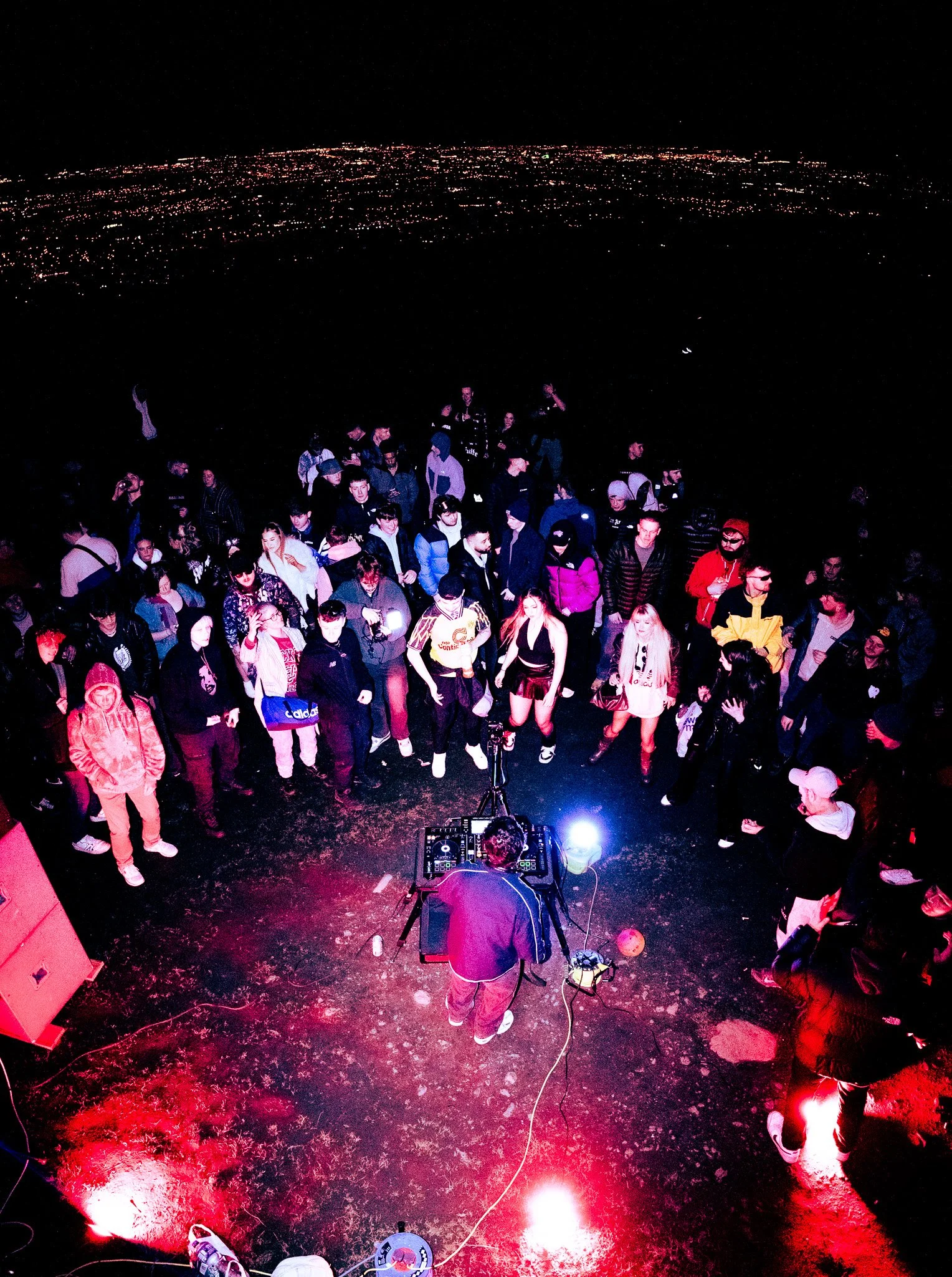 A DJ performs on a stage at night with a crowd of people dancing around him, illuminated by colorful lighting, in an outdoor setting with city lights visible in the background. Music Coverage for Irish DJ ChildsMind. 
