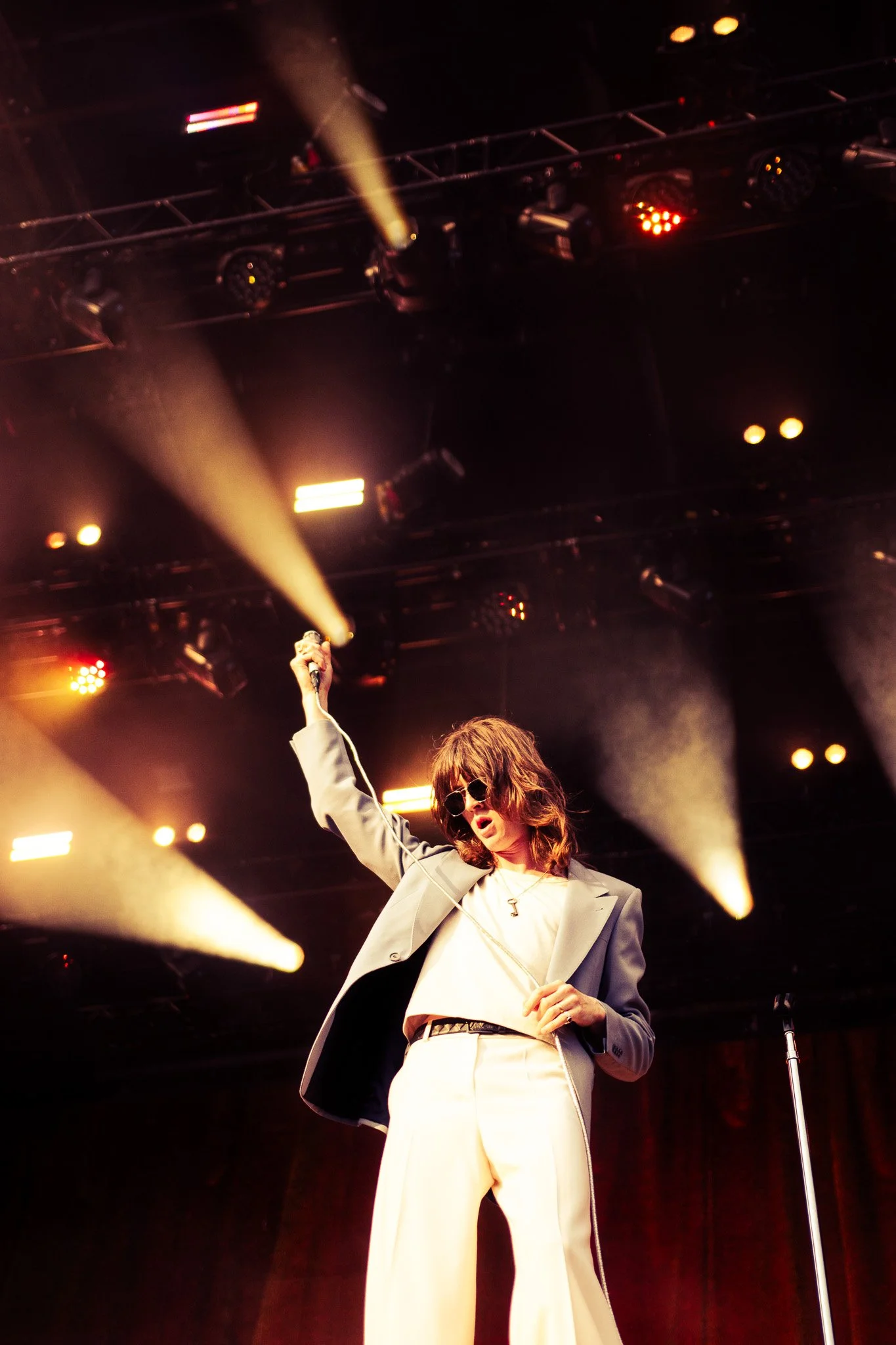 Male performer with sunglasses, brown hair, wearing a beige suit with black accents, holding a microphone in the air on stage with colorful lights and smoke. Dublin, St Annes park, Supporting Irish band Inhaler, 2025. 