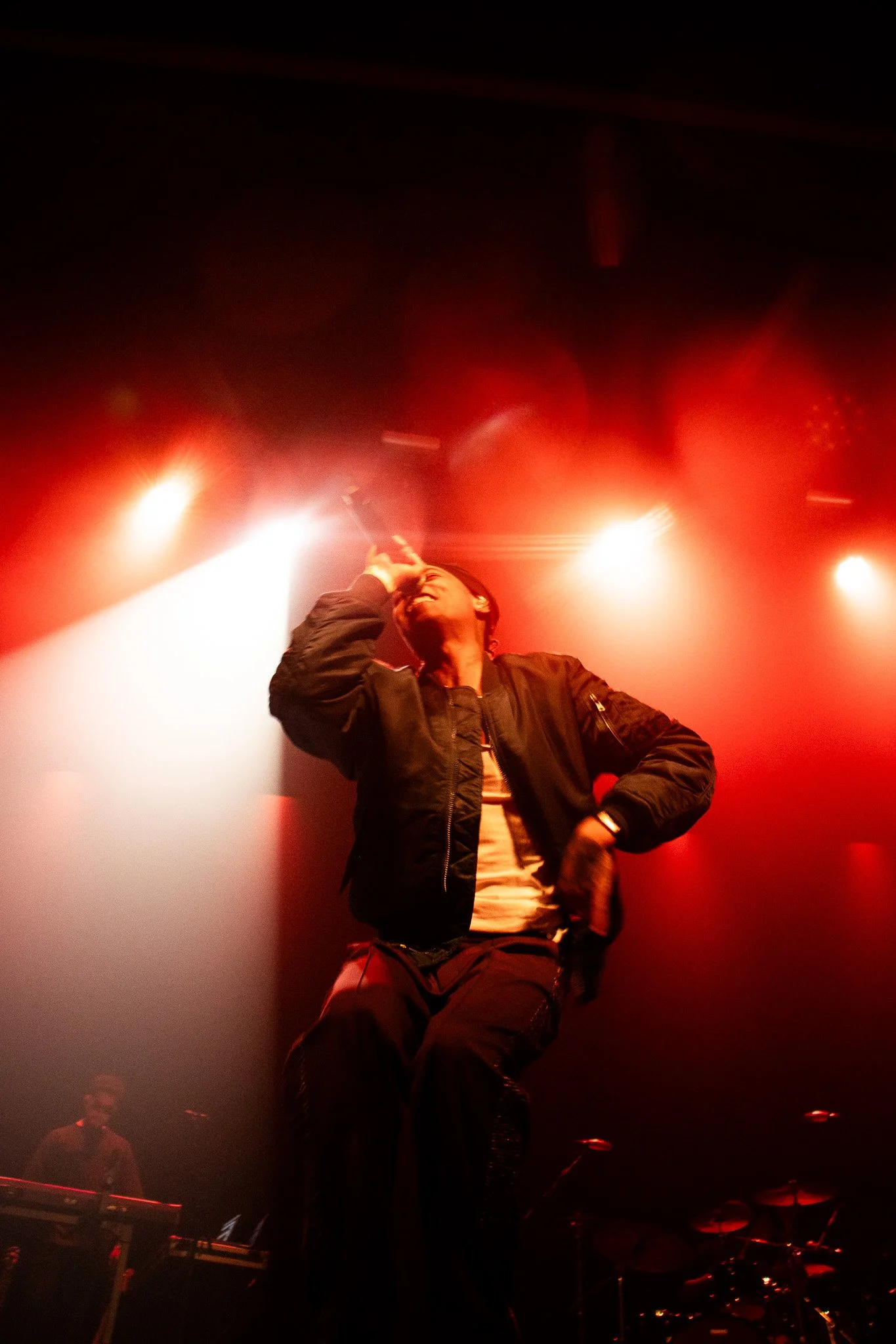Rejjie Snow in the 3Olympia Theatre 2024. Performer singing on stage under red and white stage lights, with a keyboard player in the background and drum set to the right, in a dark concert venue.
