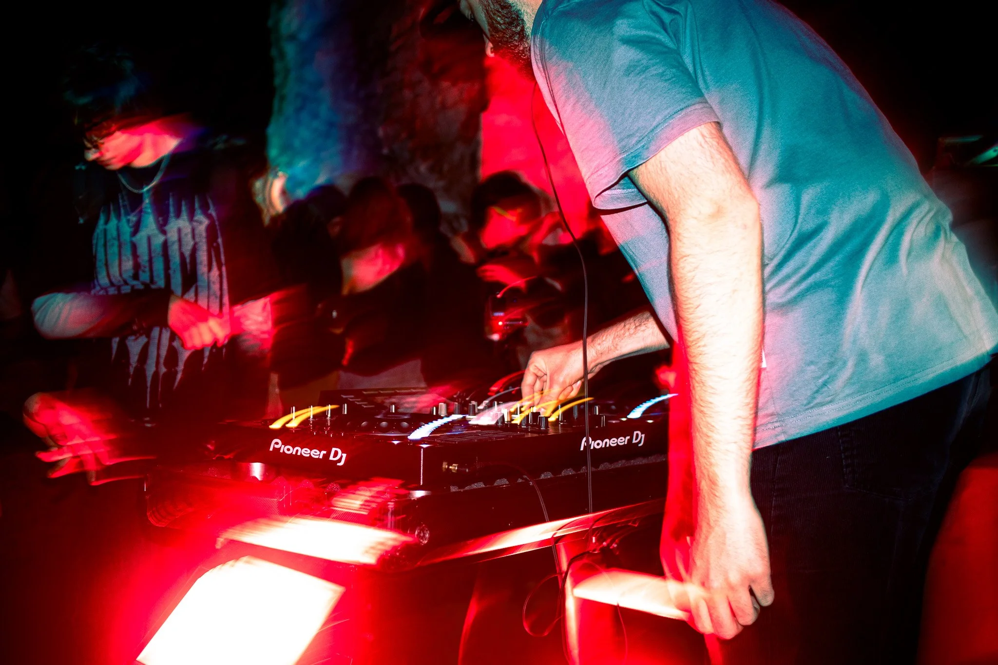 A DJ is performing at a nightclub, wearing a gray t-shirt, with people dancing in the background under red lighting.