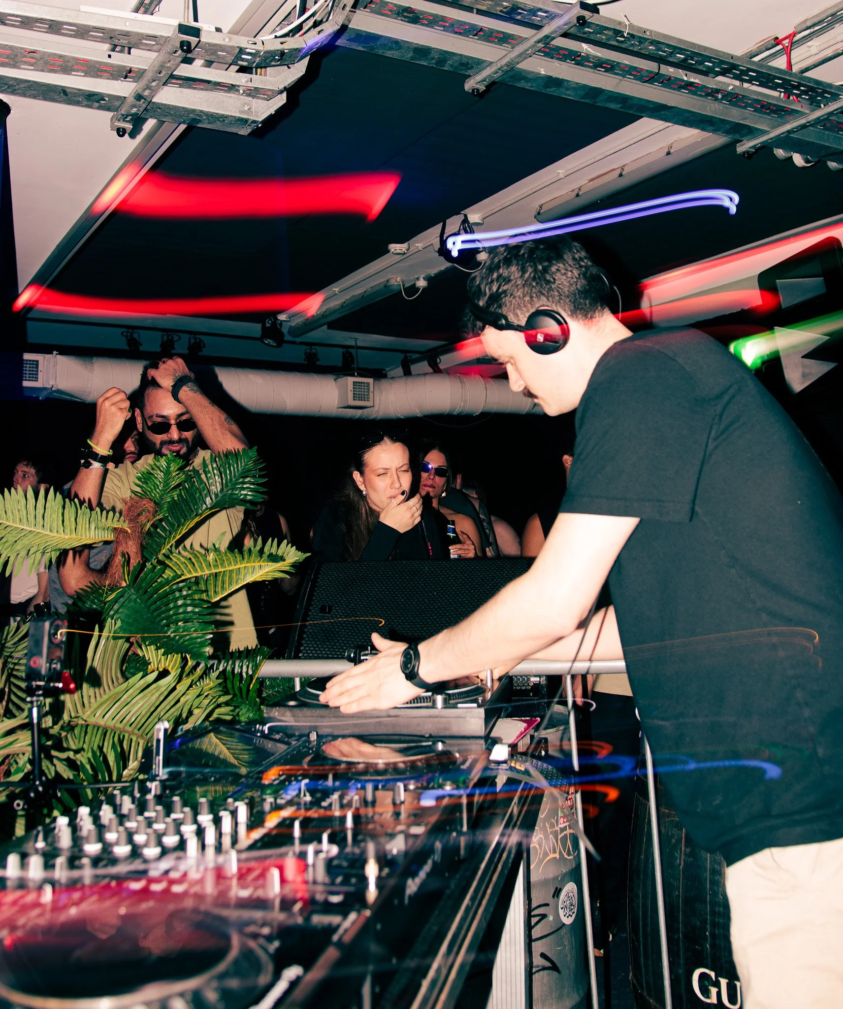DJ wearing headphones and a black T-shirt performing at a nightclub with people dancing and enjoying the music.