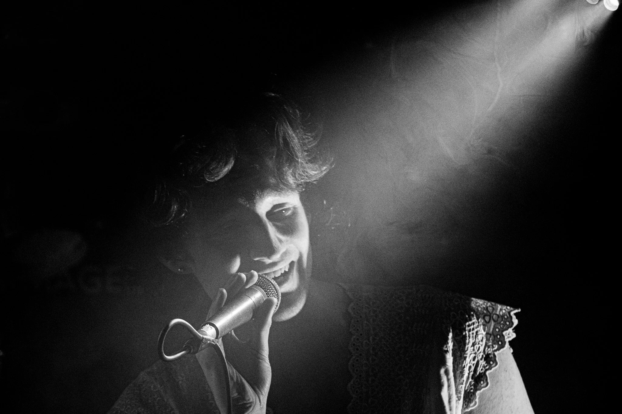 Black and white photo of a man singing into a microphone, with a light shining on her face, wearing a lace top.