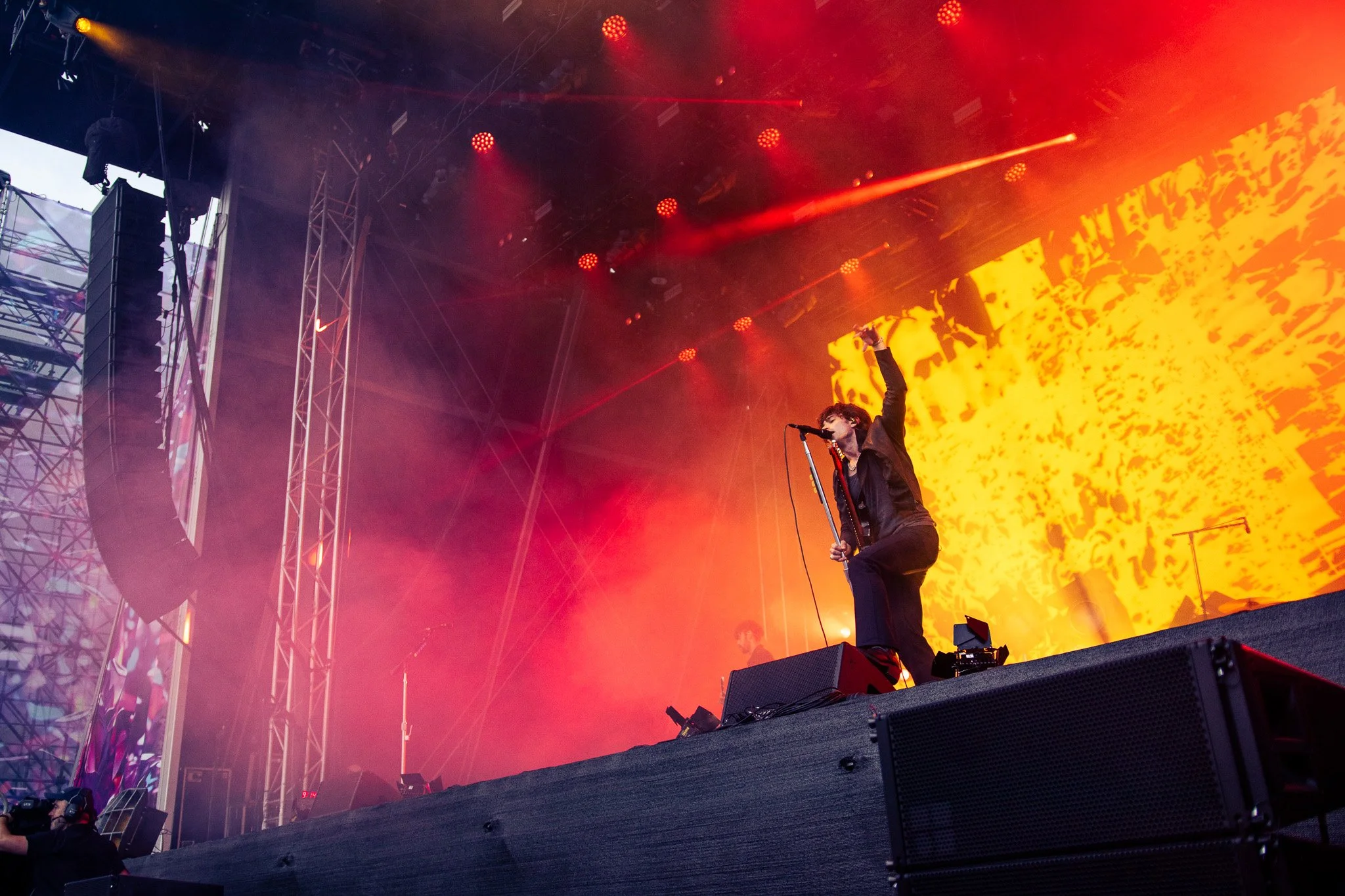 A singer performing on an outdoor stage with orange and yellow abstract background, surrounded by metal trusses and lighting, with a person in the audience capturing the moment. Dublin, St Annes park, Irish band Inhaler, 2025. Music Photography
