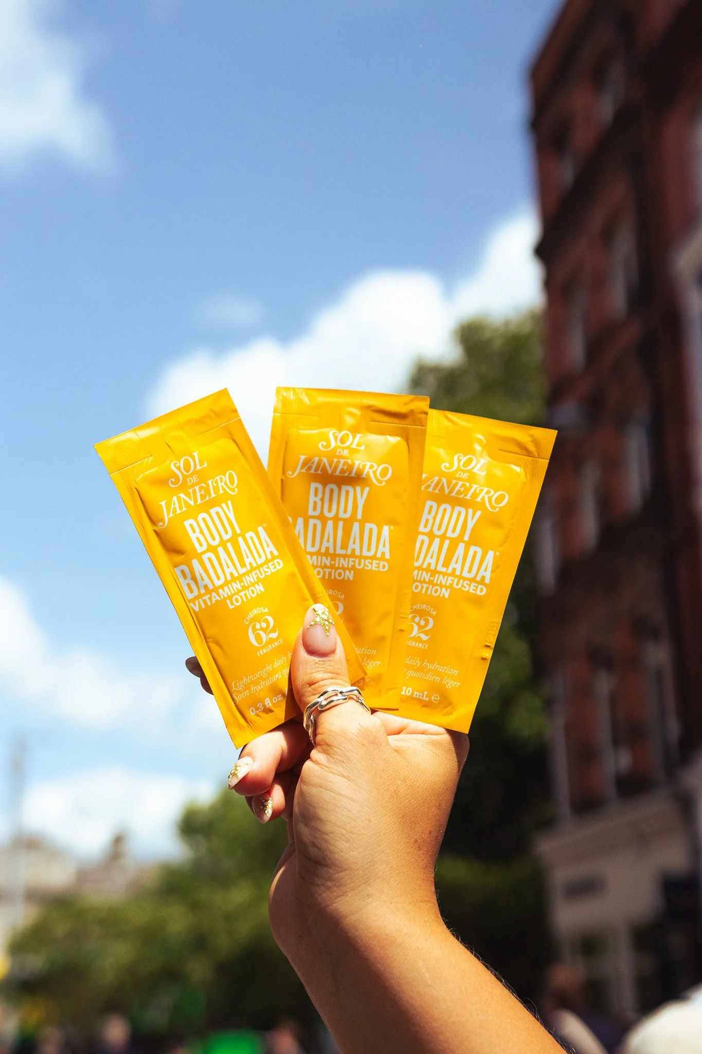 A hand holds three yellow packet sachets of Sol de Janeiro Body Badalada vitamin-infused lotion with a bright blue sky and a building in the background.