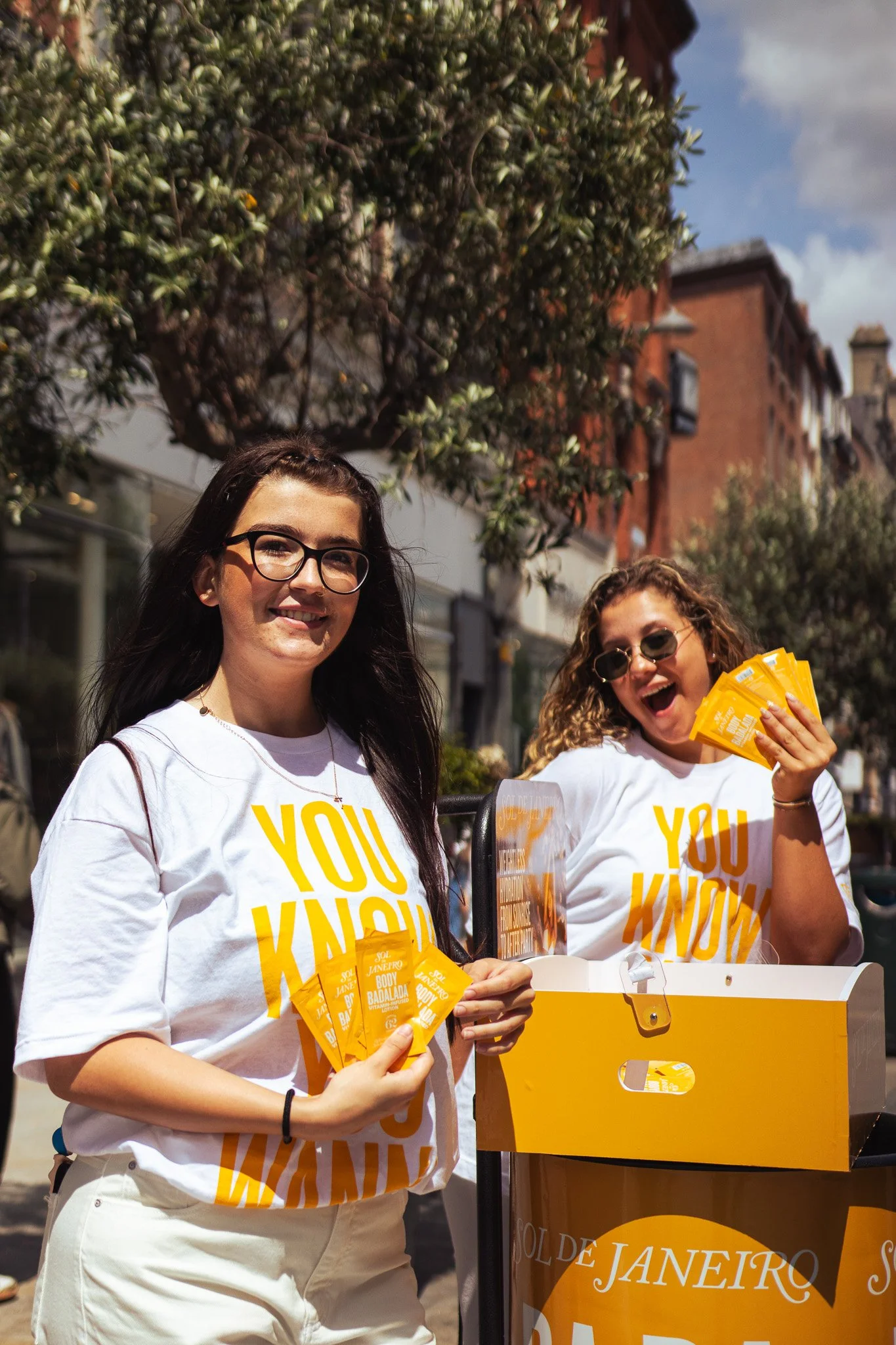 Two women promoting Sol de Janeiro skincare products outdoors, holding yellow samples, wearing white T-shirts with yellow text, smiling. On Street Brand activation photography coverage. 