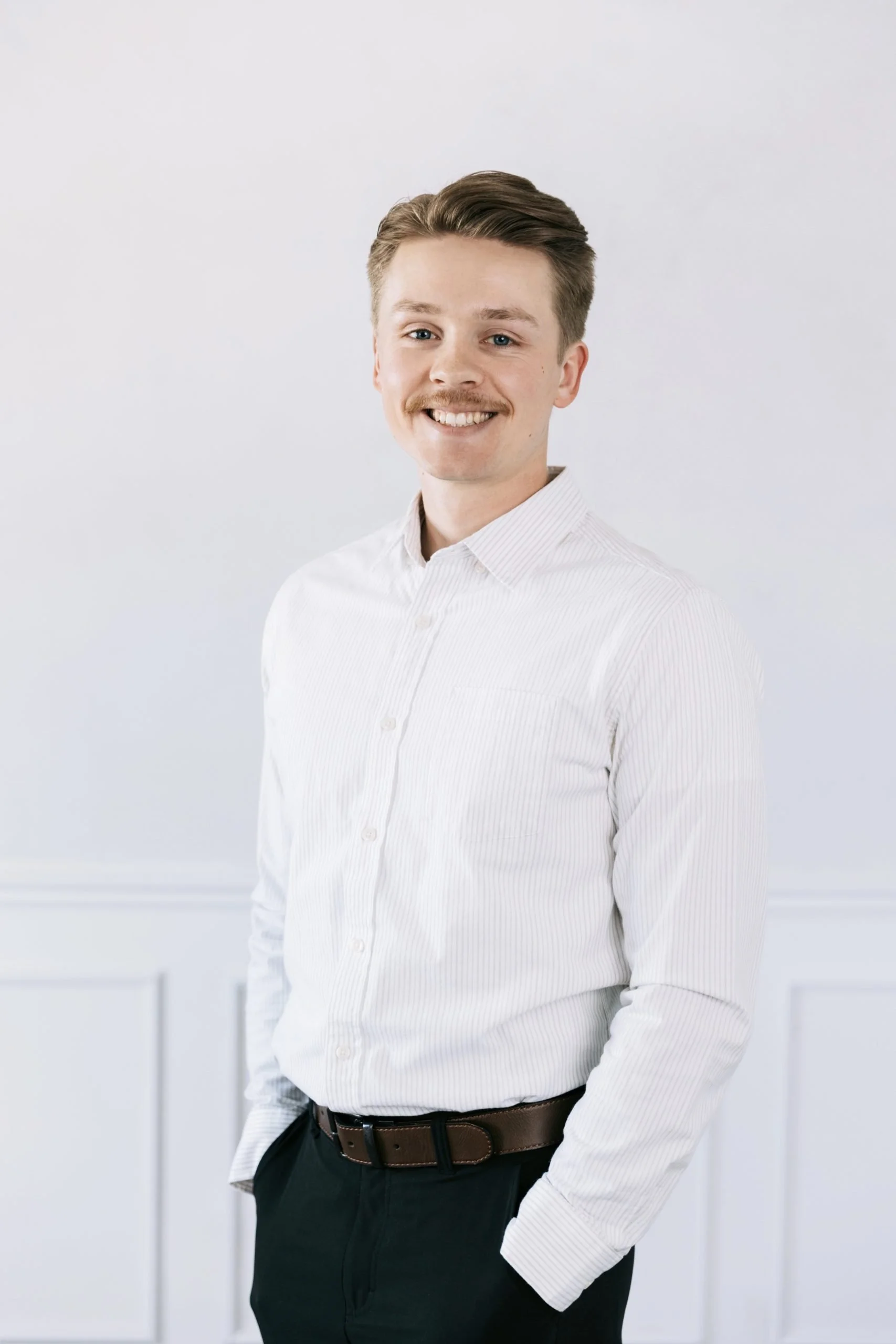 A young man with light skin, brown hair, and a mustache is smiling at the camera. He is wearing a white, long-sleeved, button-up shirt and dark trousers, standing against a plain light-colored background.