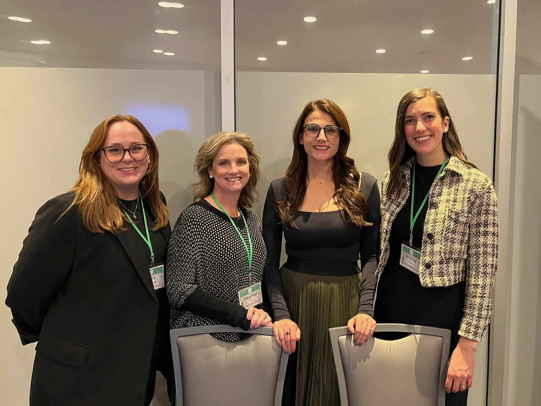 Four women standing together at a conference, wearing name tags on green lanyards, with a glass wall behind them.