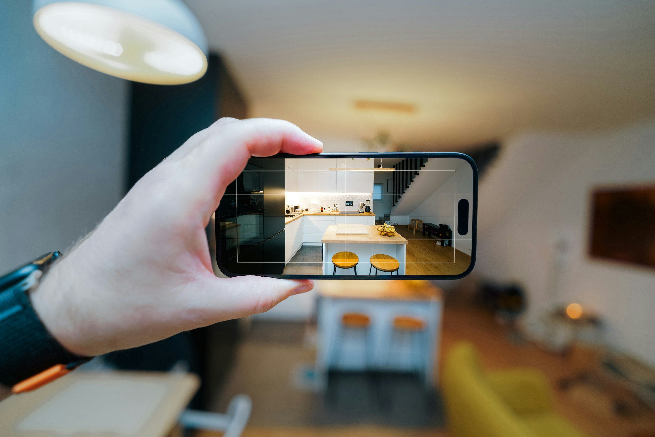 A person holding a smartphone, taking a photo of a modern kitchen with white cabinets, a kitchen island, two yellow barstools, and a staircase in the background.