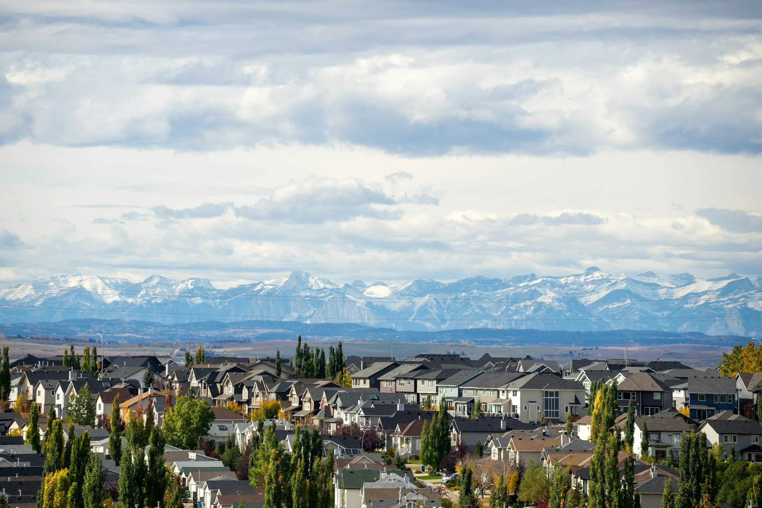A suburban town with closely packed houses, trees surrounding the neighborhood, and snow-capped mountains in the background under a cloudy sky.