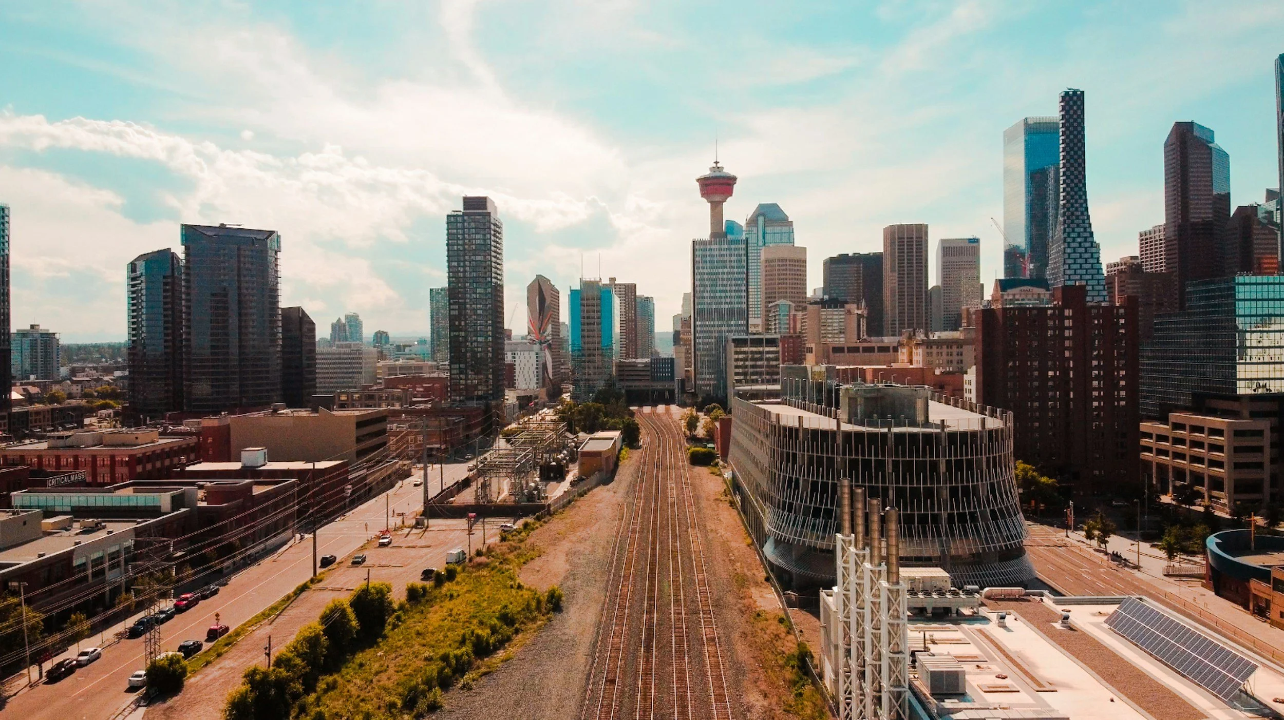 A city skyline with tall skyscrapers, a tower with a red top, railway tracks, and a modern building with a curved facade on a clear day.