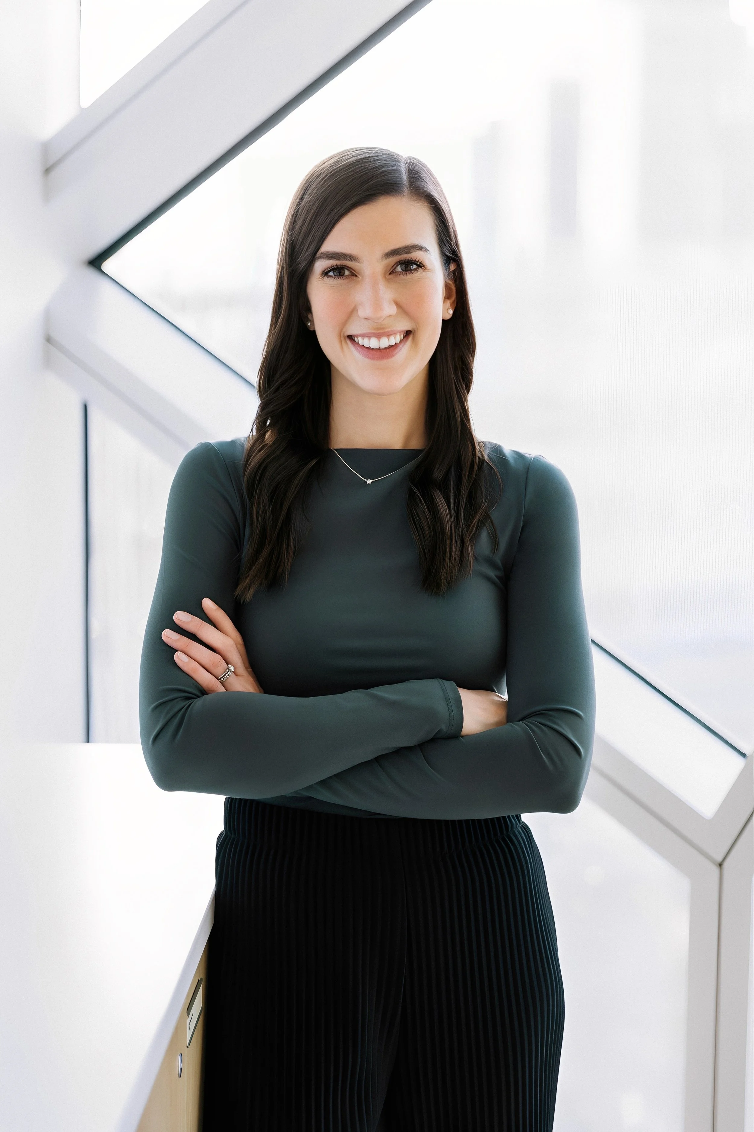 A woman with long dark hair smiling, standing with arms crossed in front of a white modern architectural background with angled windows.