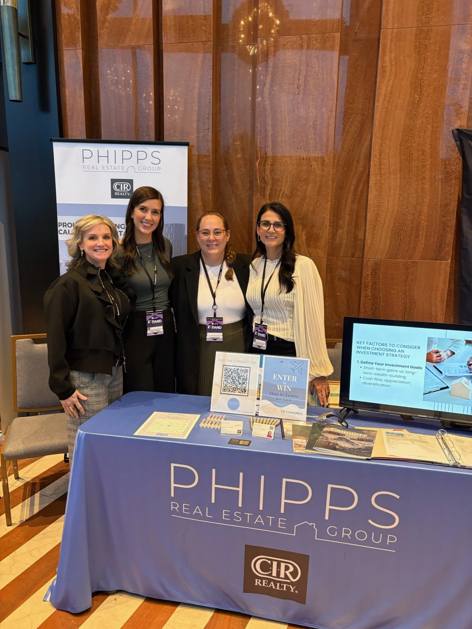 Four women standing behind a promotional table for PHIPPS Real Estate Group at a conference, smiling for the camera. The table has a blue tablecloth with the company logo, various informational materials, a QR code sign, and a monitor displaying key factors for investment strategy.