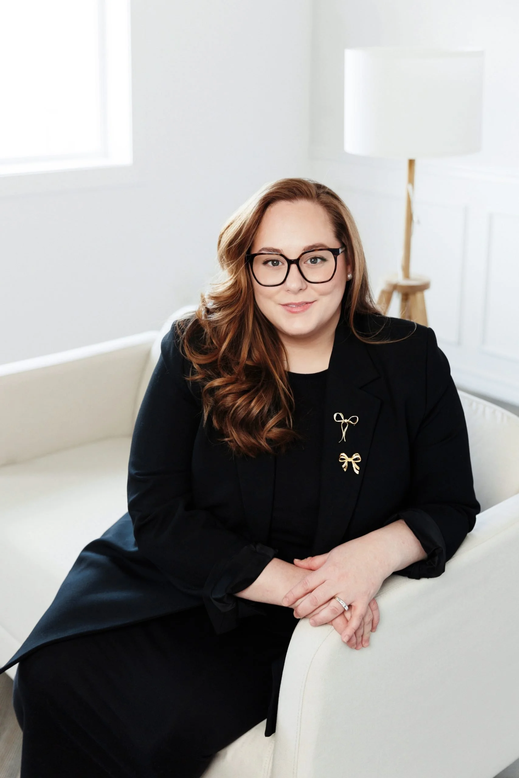 A woman with long red hair, glasses, and a black blazer with bow pins sitting on a white sofa in a bright, white room.