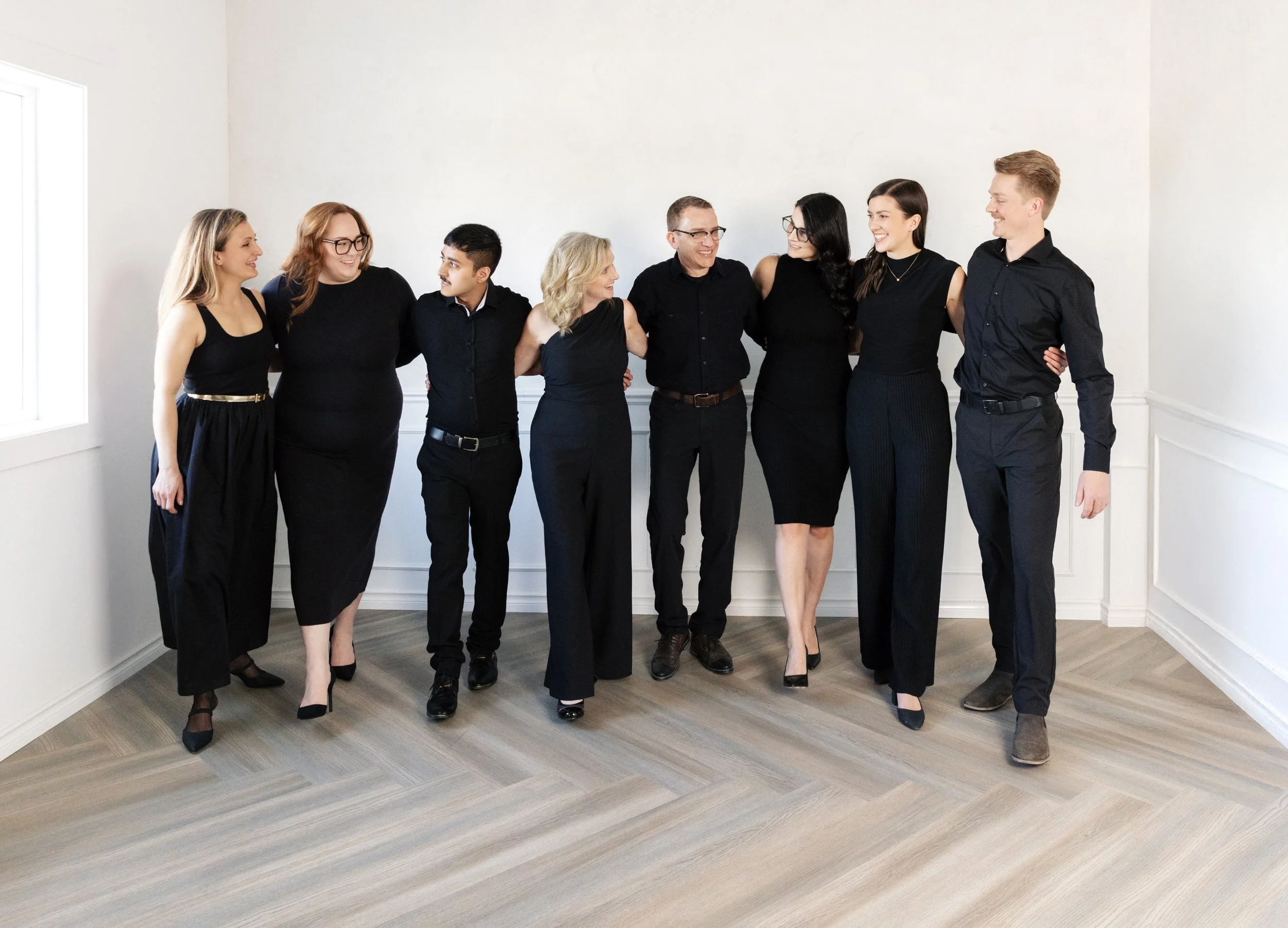 Group of eight diverse people standing arm-in-arm in a room, dressed in black formal clothing, smiling, celebrating.