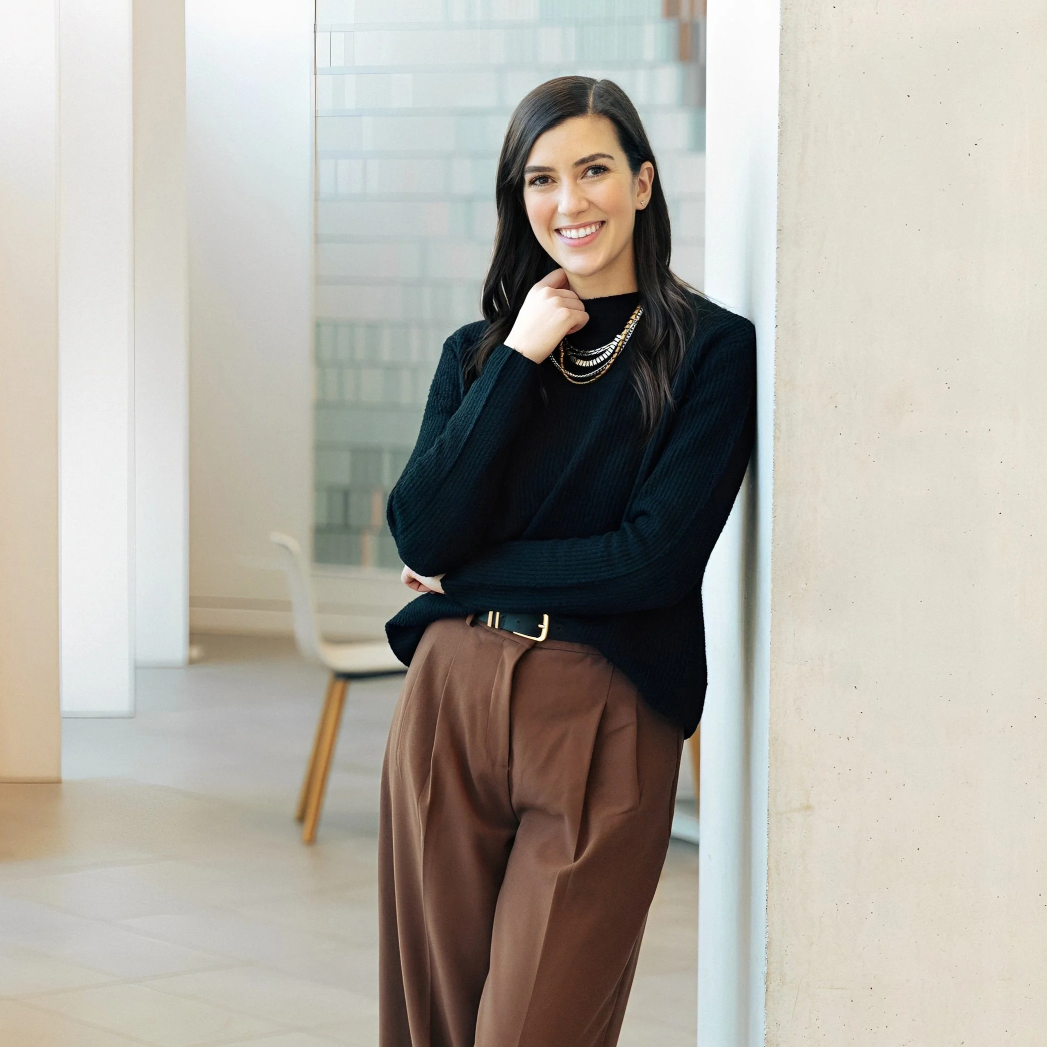 A woman smiling, wearing a black sweater and brown pants, leaning against a wall in a modern indoor space.