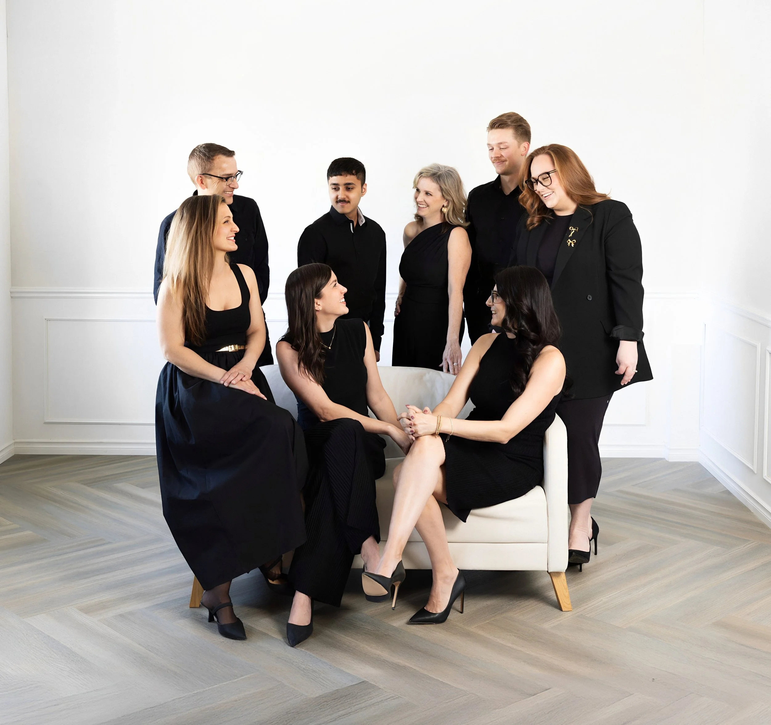 Group of nine diverse people dressed in black, posed in a bright, white room with wooden flooring, some sitting on a sofa and others standing around, smiling and engaging with each other.